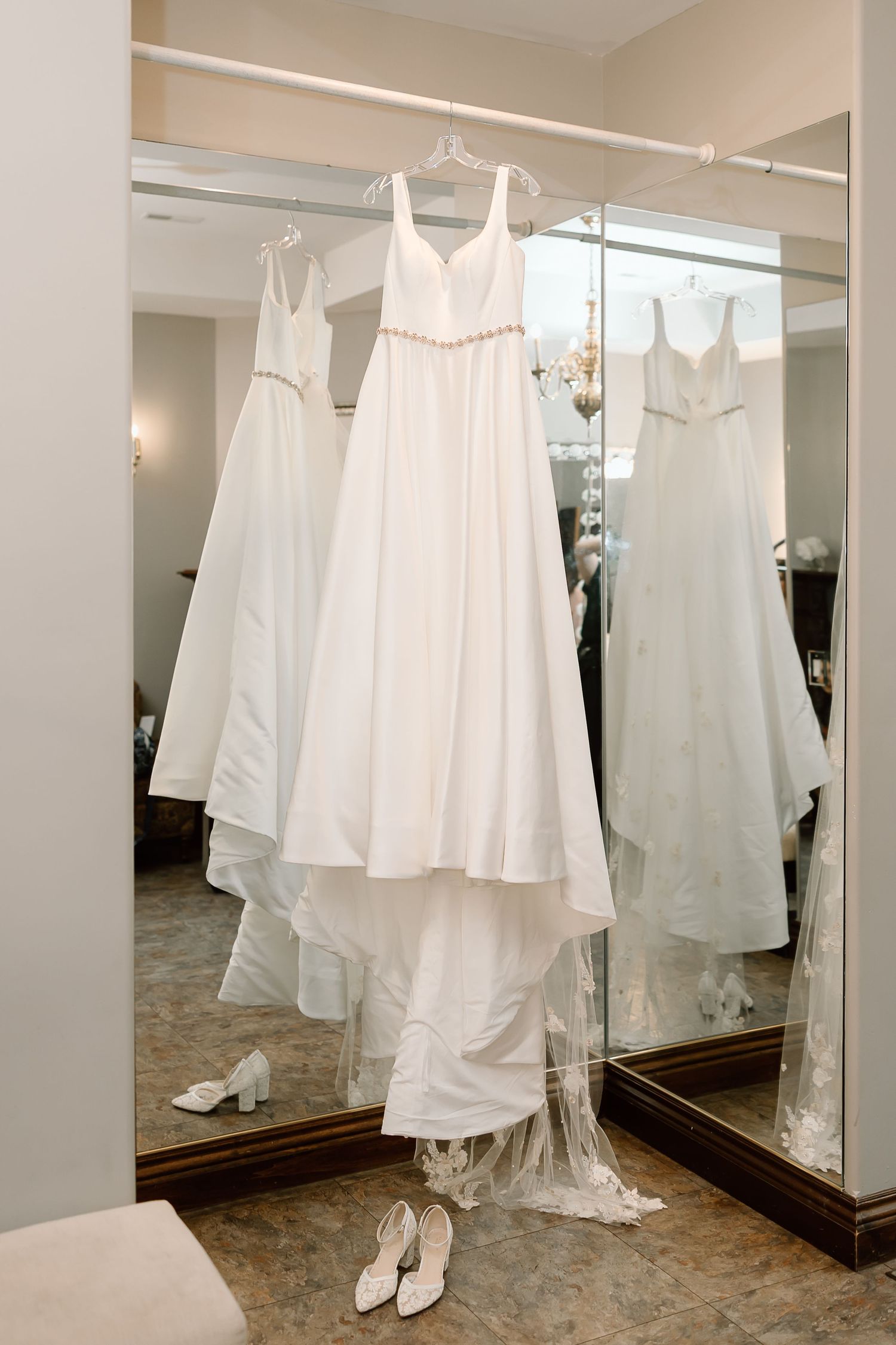 White wedding dress hanging in bridal boutique fitting room with mirrors and white shoes on floor.