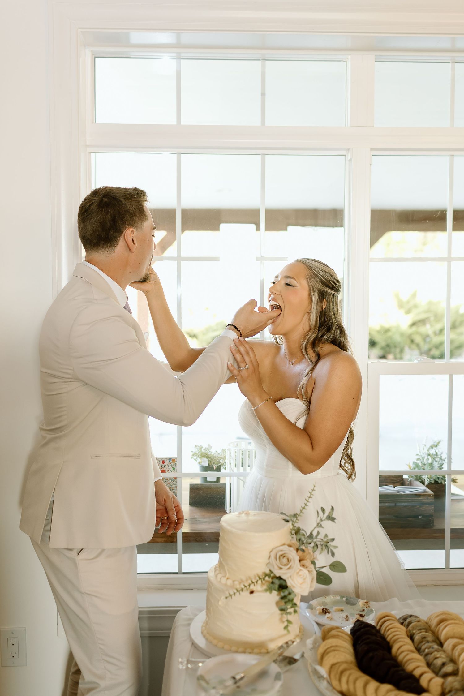 Newlyweds playfully feed each other wedding cake at their reception by a bright window.