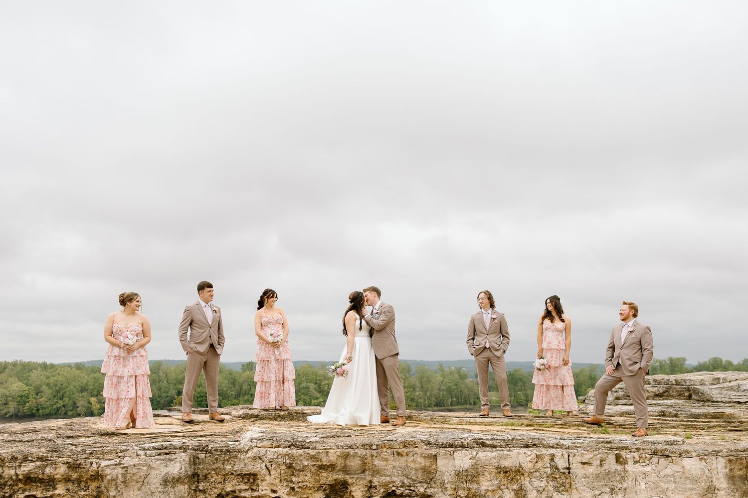 Wedding party in pink and tan attire poses on rocky outcrop under cloudy sky.