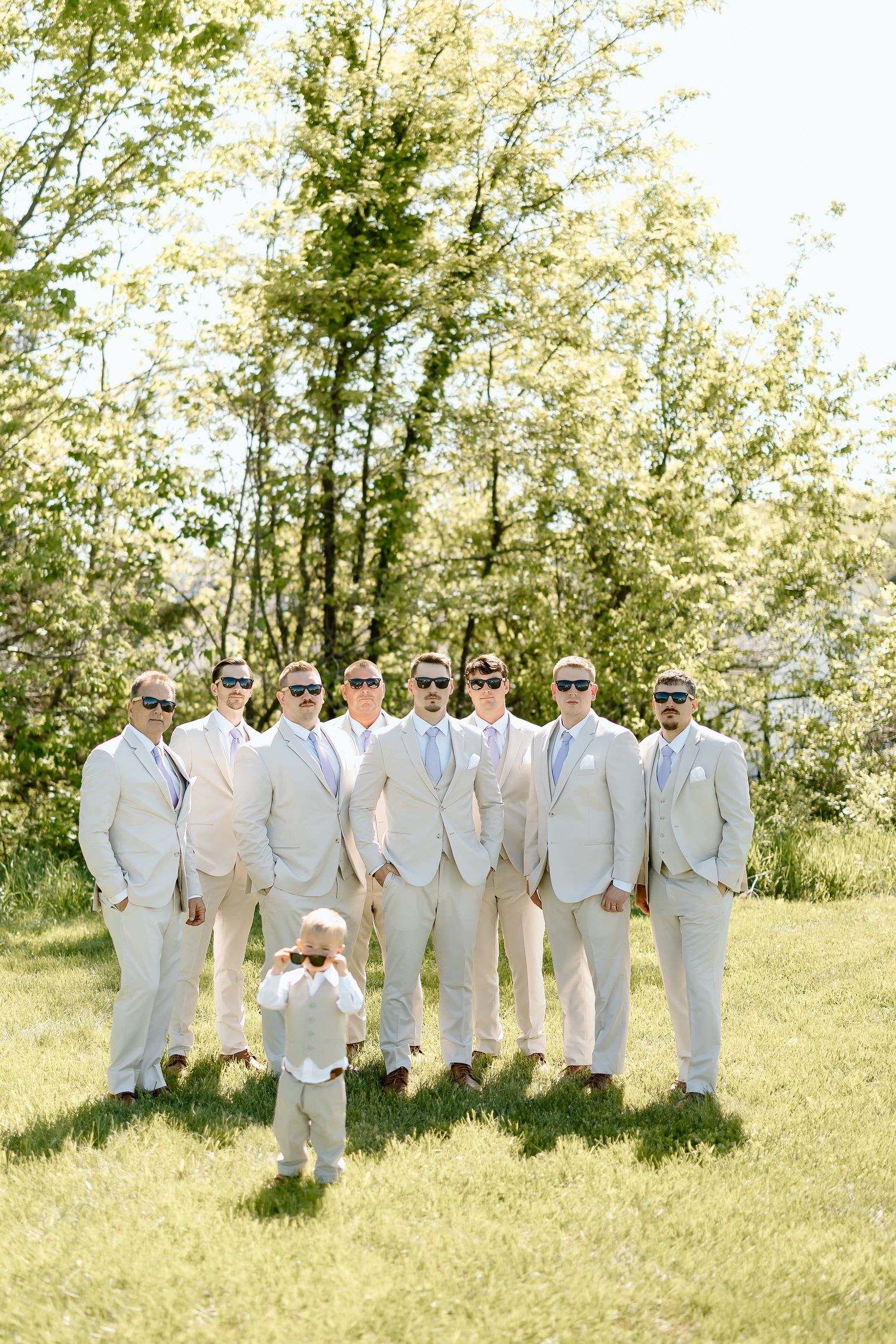 Groomsmen in light beige suits pose together outdoors with a young ring bearer in front on a sunny day.