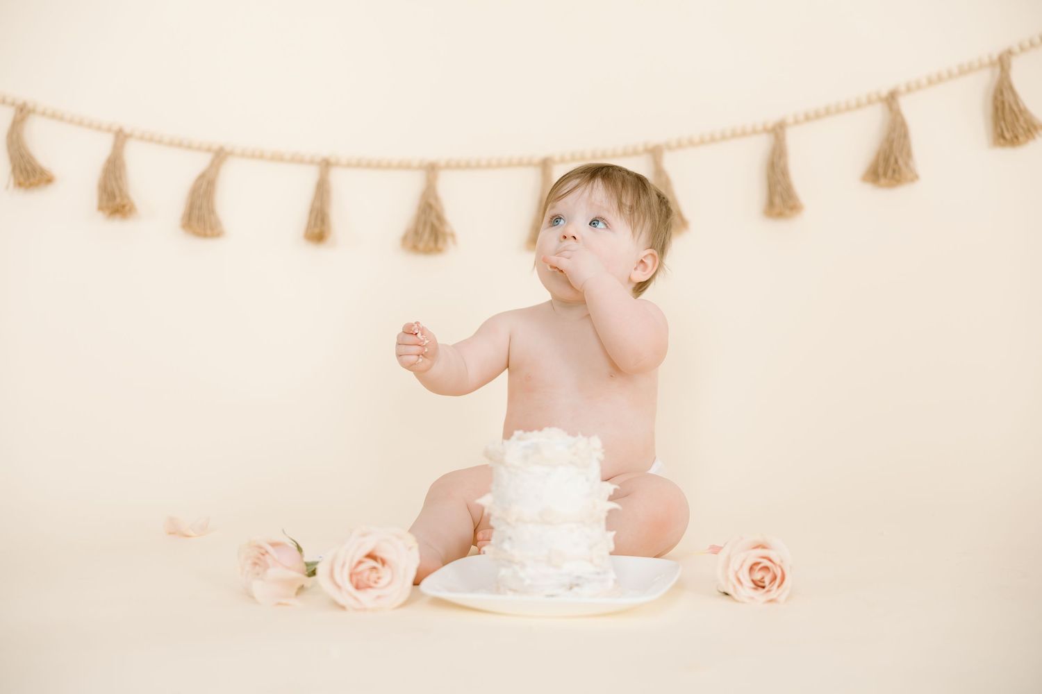 A baby reaches up while sitting with a white cake and pink roses against beige backdrop.