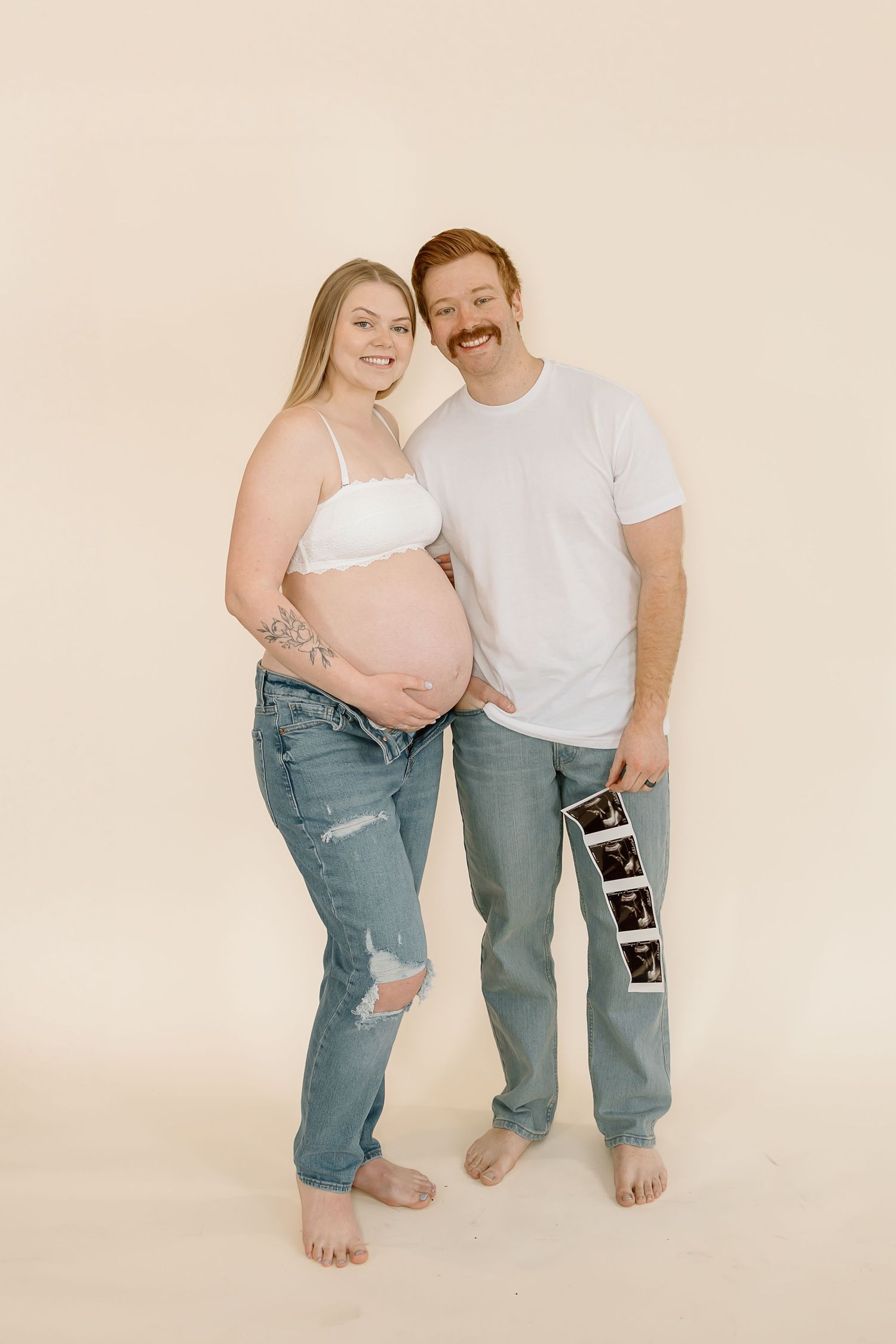 A barefoot couple poses together for a pregnancy announcement photo against a neutral backdrop.