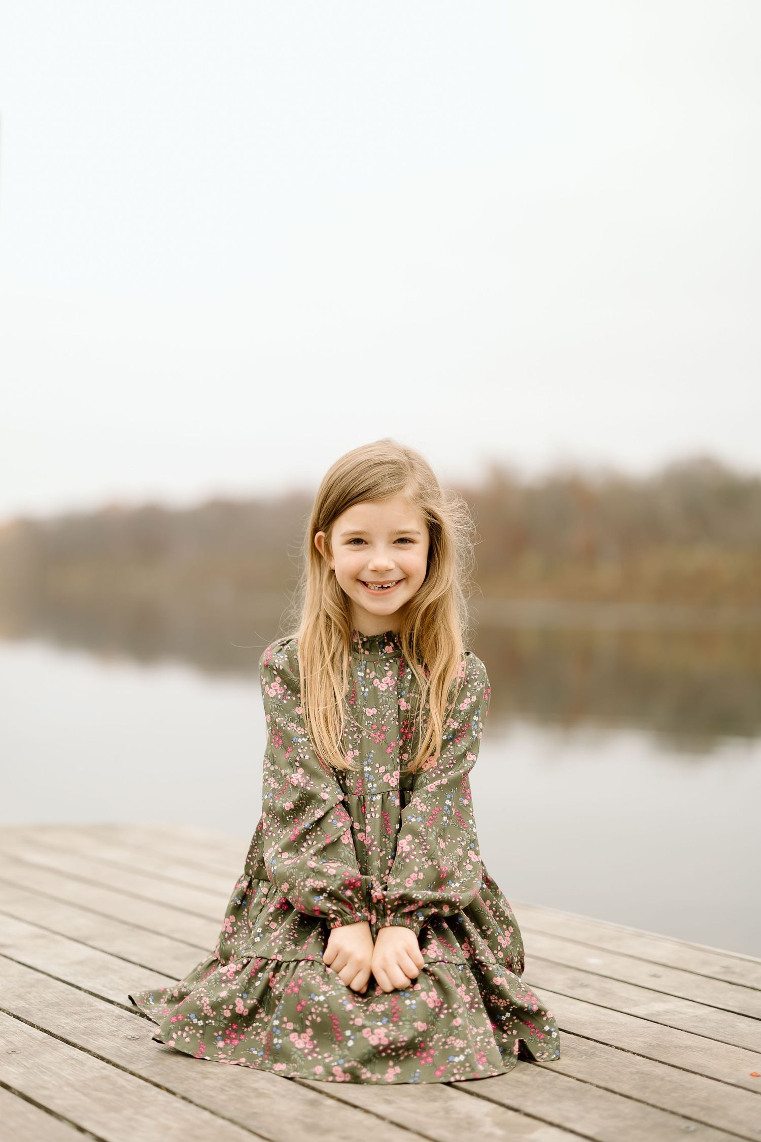 Smiling child in floral dress sits on wooden dock during lakeside photo session.