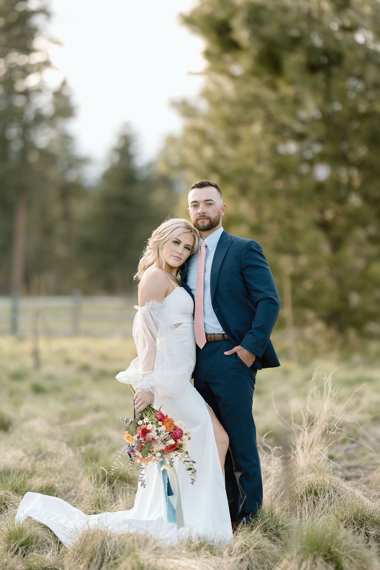 Bride and groom pose together in rustic field with mountains and forest in background.