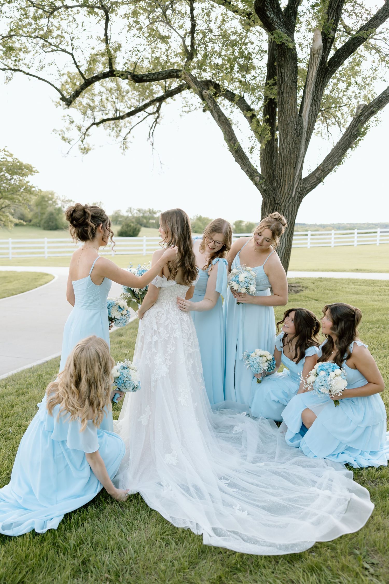 Bride in white lace gown surrounded by bridesmaids in light blue dresses under a large tree outdoors.