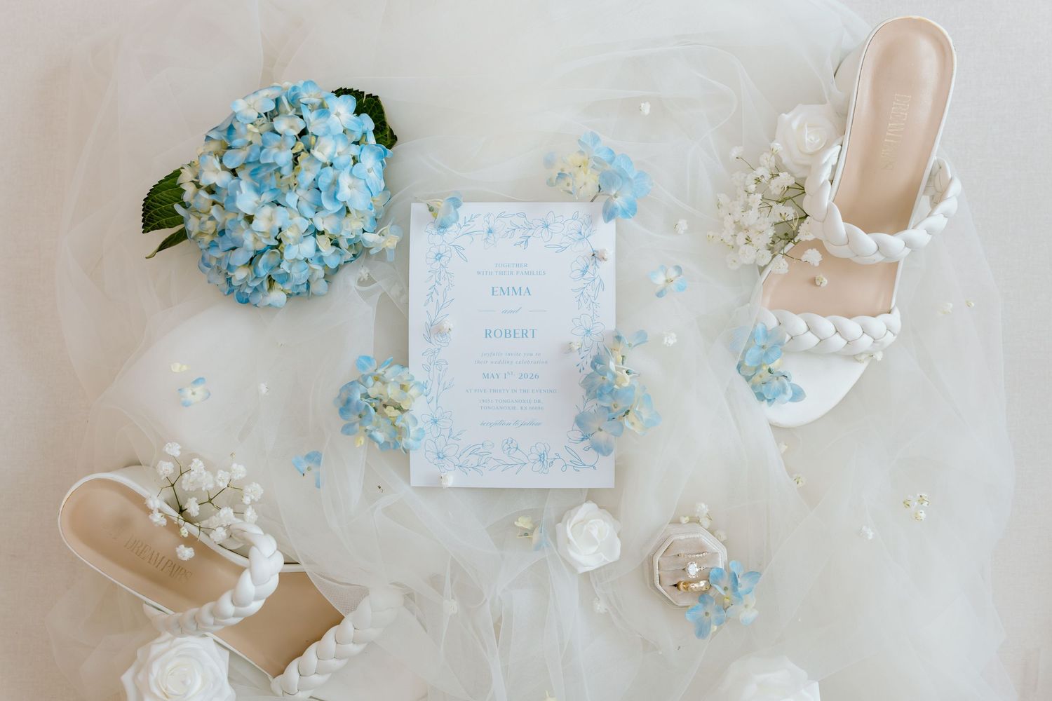 Blue hydrangea wedding flat lay with invitation, pearl heeled sandals, and floral accents on white tulle.