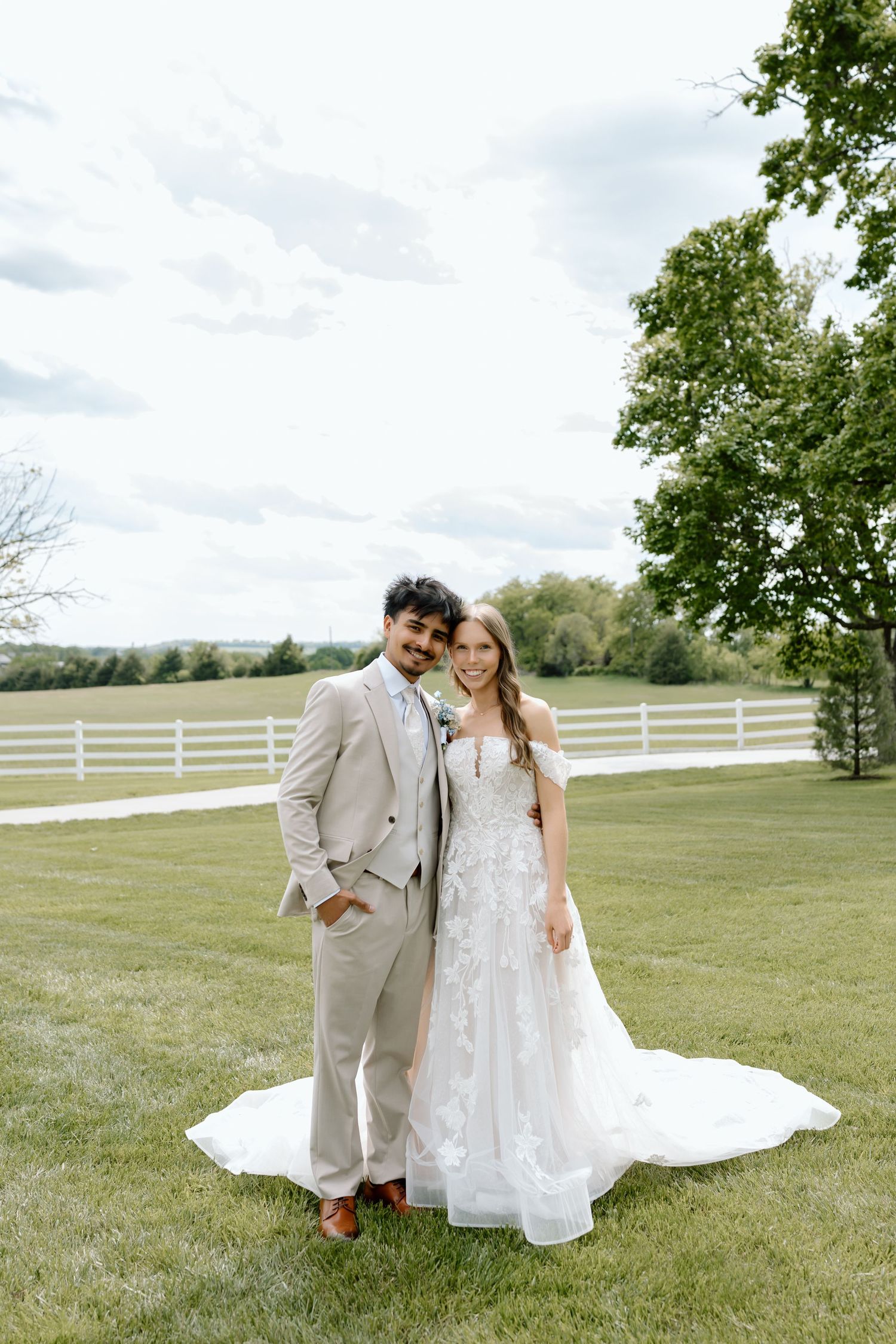 A bride and groom pose outdoors on green grass, she in a floral gown and he in a tan suit, at a farm venue.