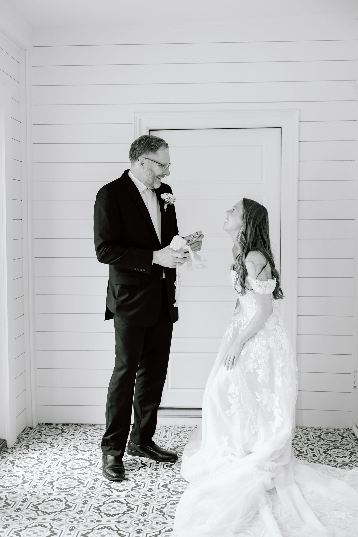 Black and white photo of a bride in lace gown sharing an emotional moment with a man in a suit reading a note.