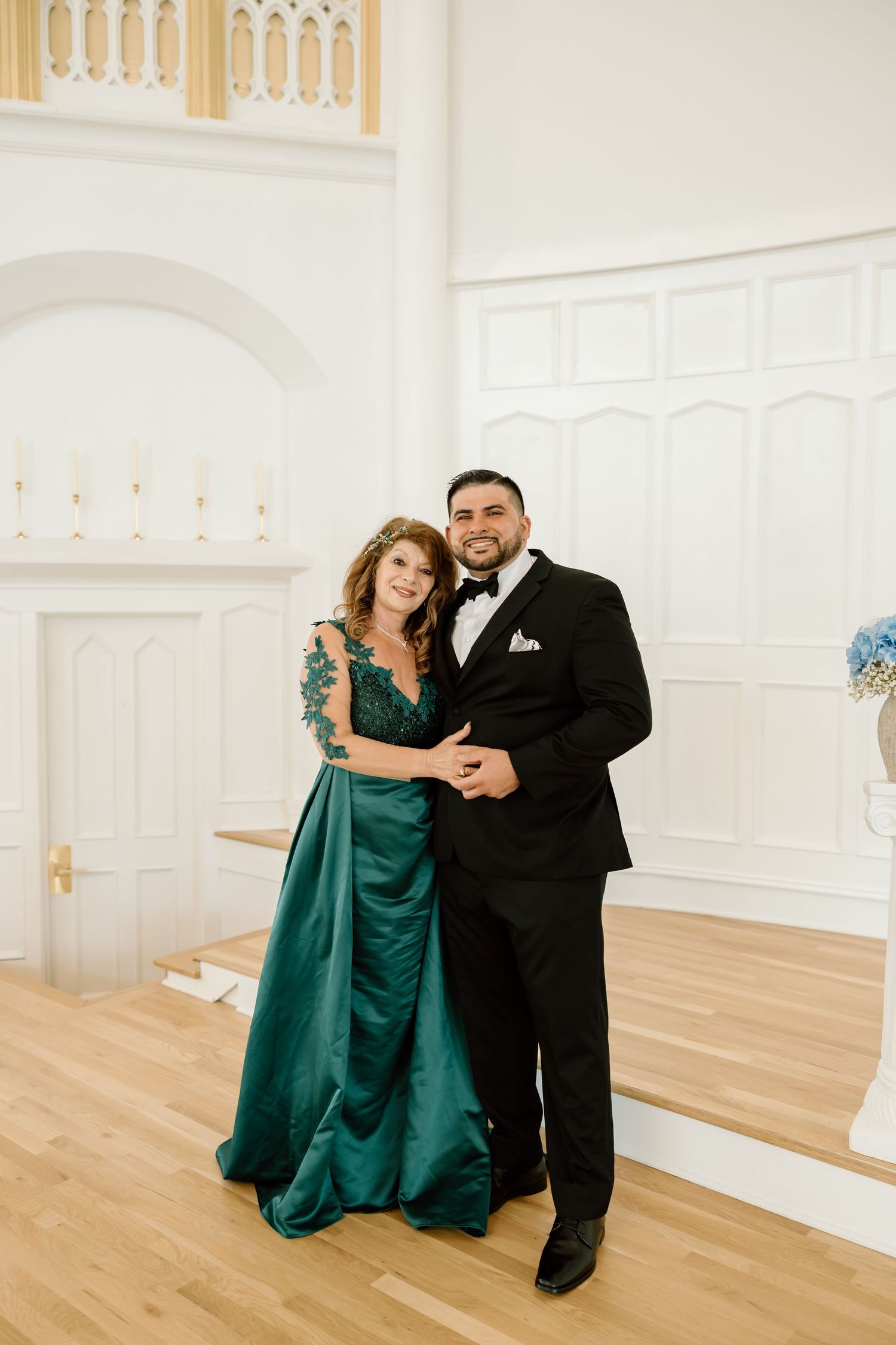 Couple posing in elegant formalwear, woman in green gown and man in black tuxedo, inside a white wedding venue.