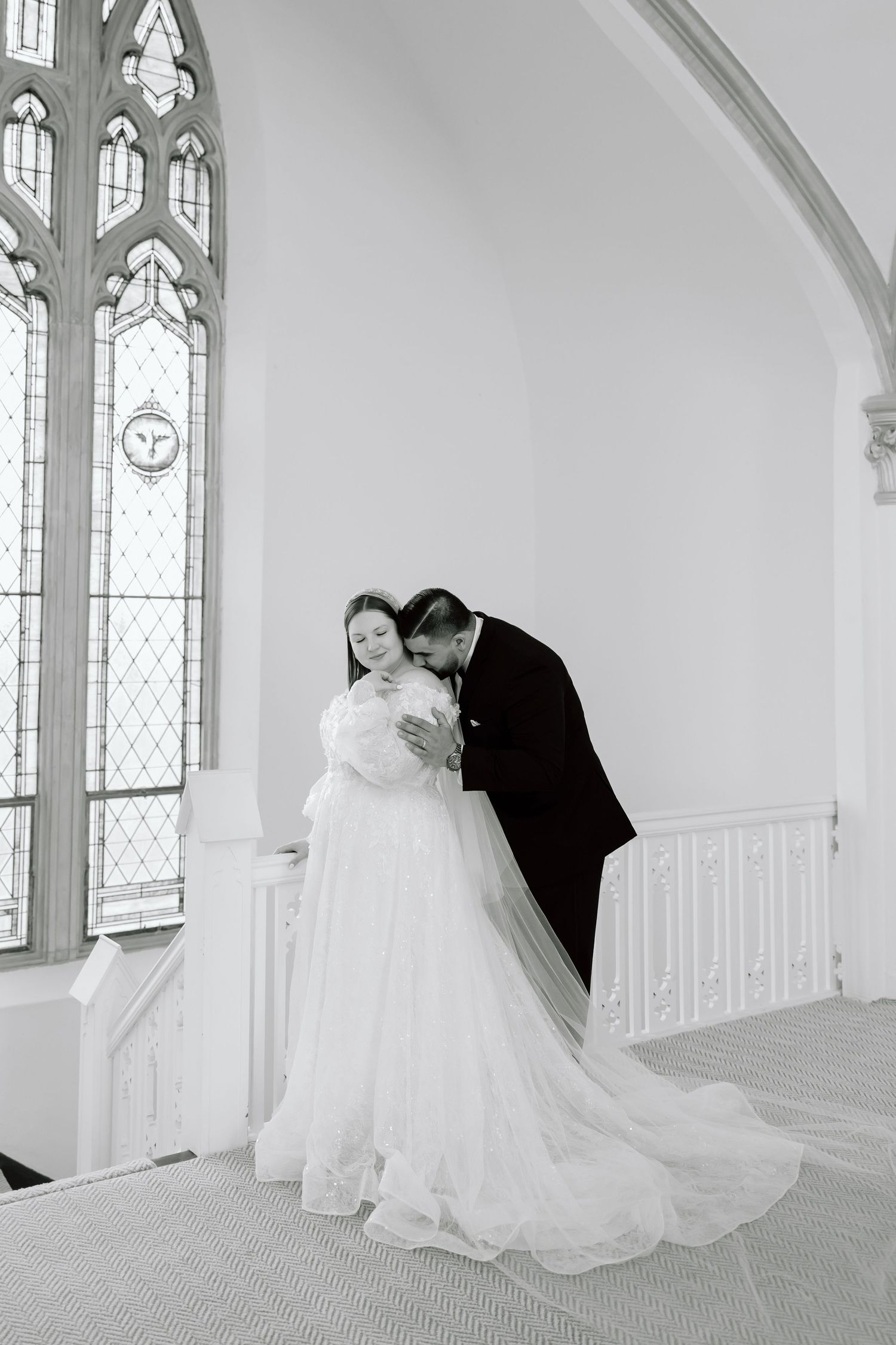 Black and white wedding photo of bride and groom sharing tender moment in elegant church with gothic stained glass windows.