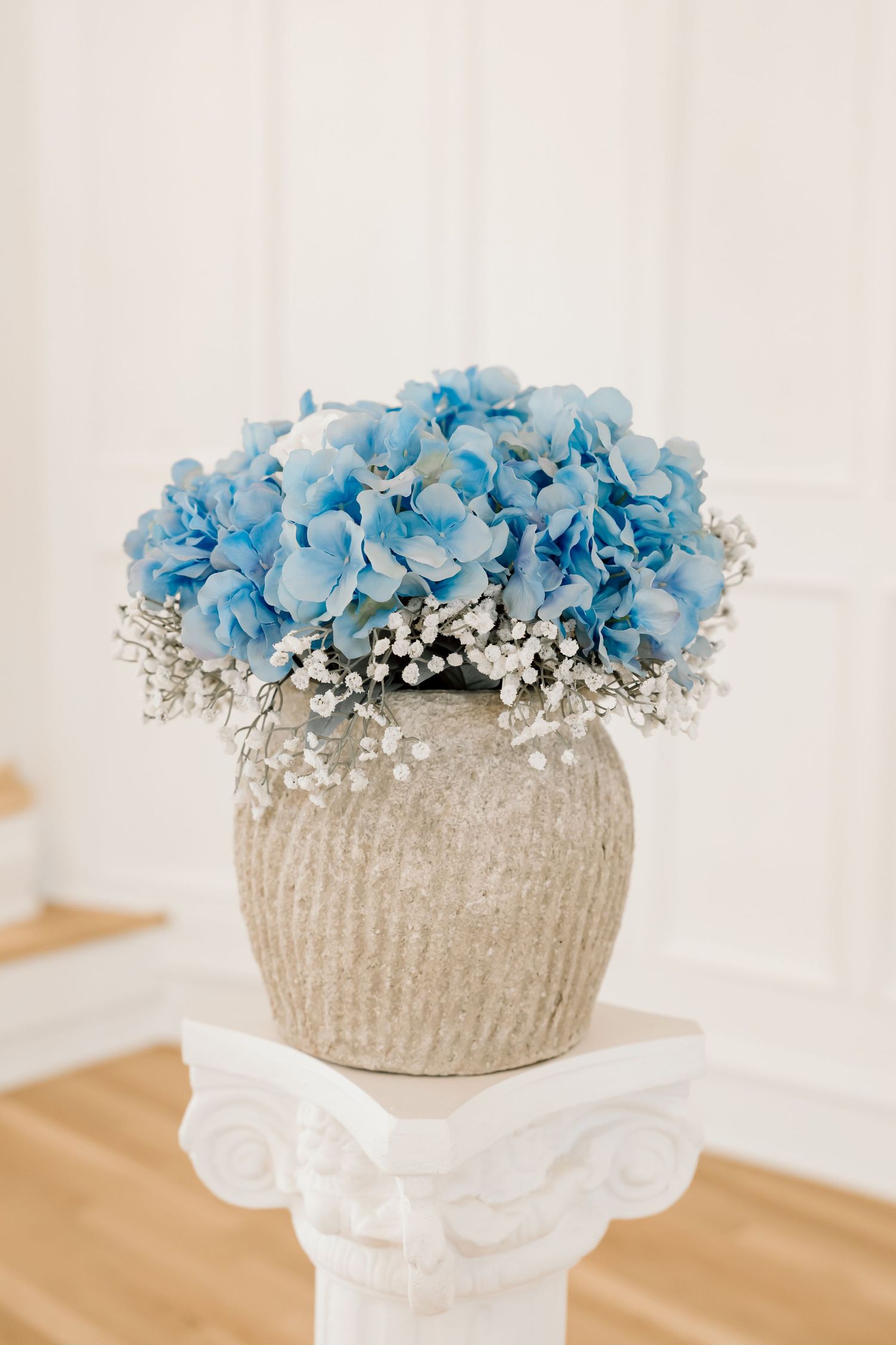 Blue hydrangeas and white baby's breath arranged in a rustic stone vase on a white decorative pedestal.