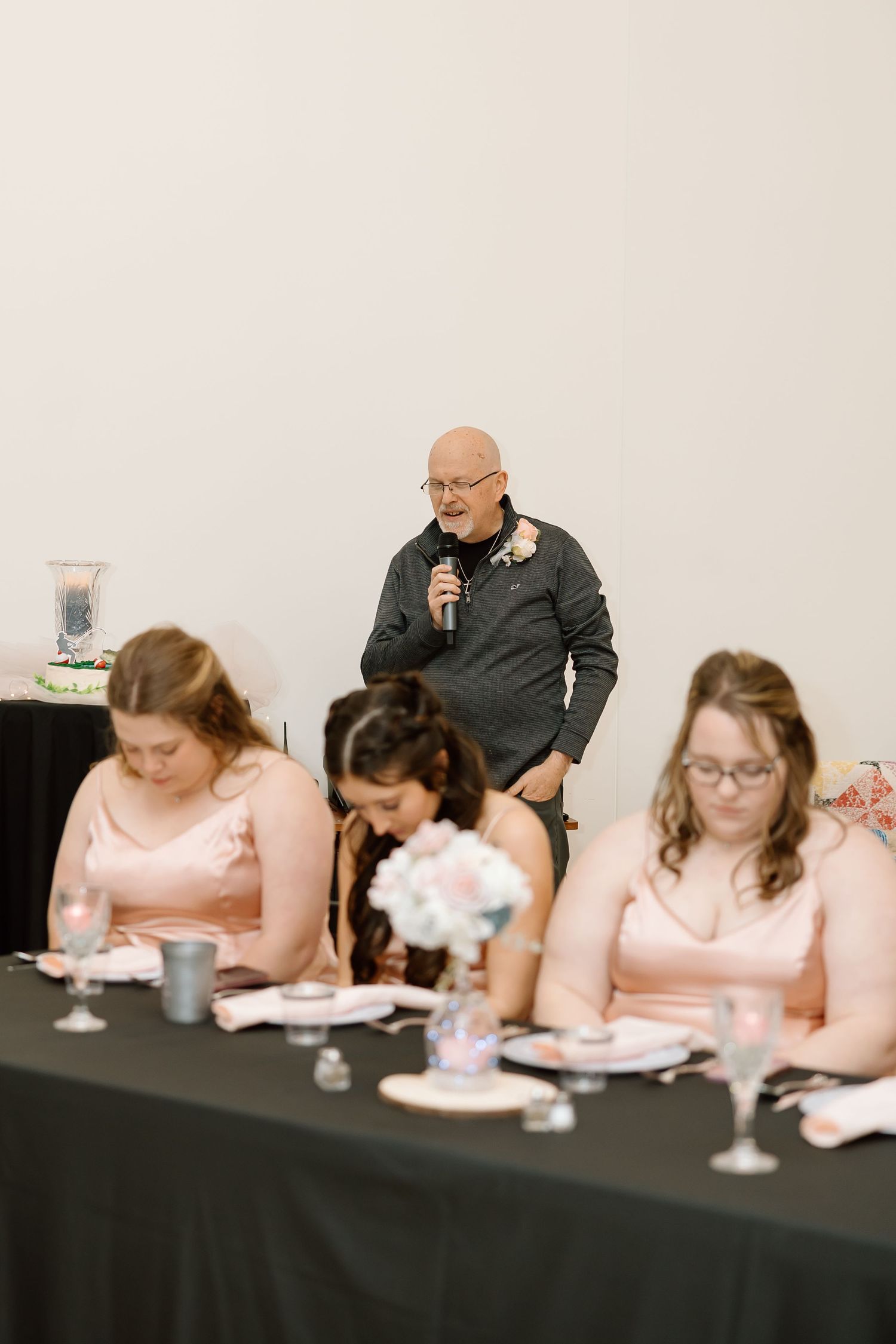 Someone speaks into a microphone while standing behind a table where others in pink dresses are seated at a formal event.