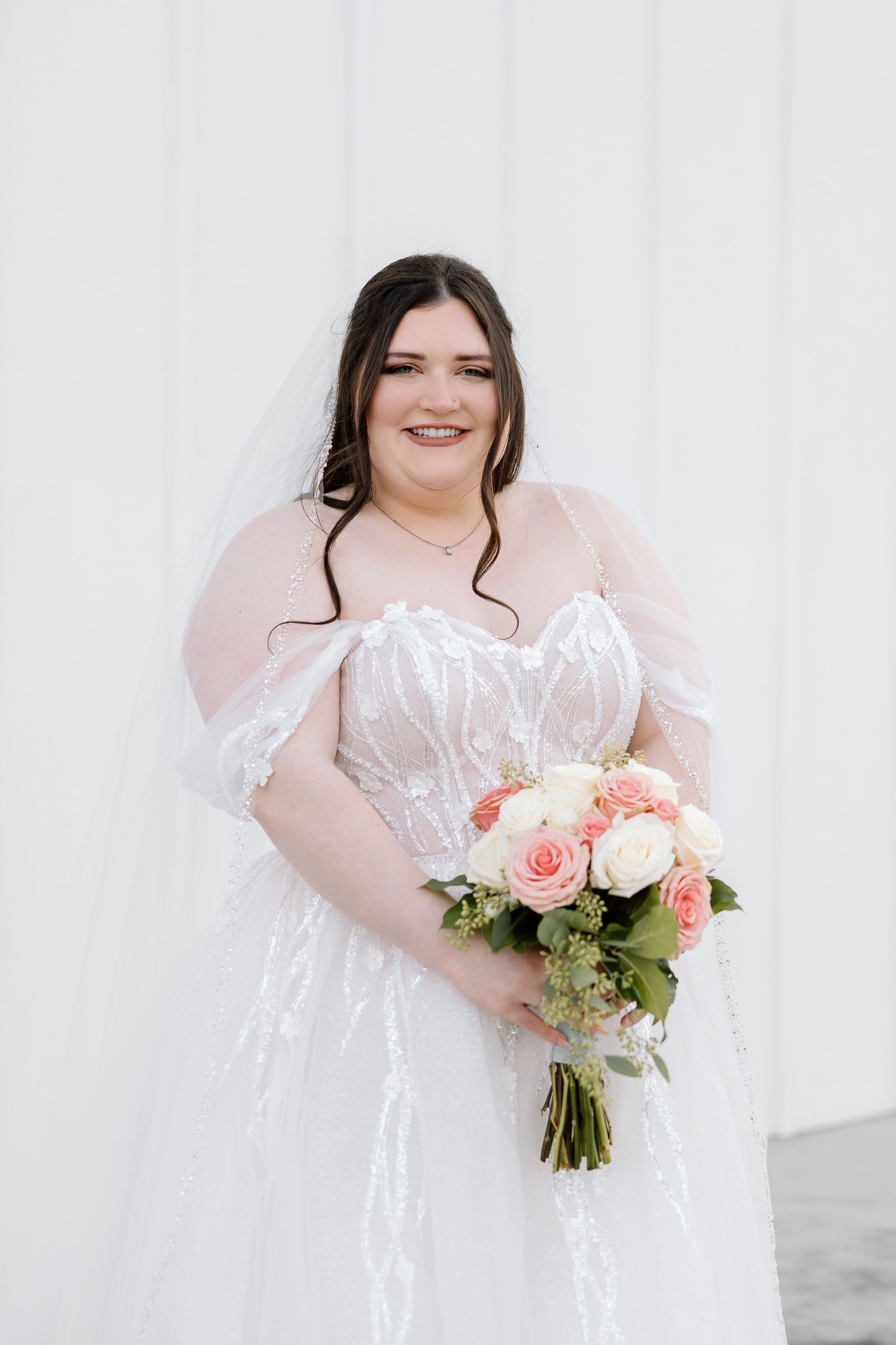 A bride in a white lace gown holds a beautiful bouquet of pink and white roses against a white backdrop.