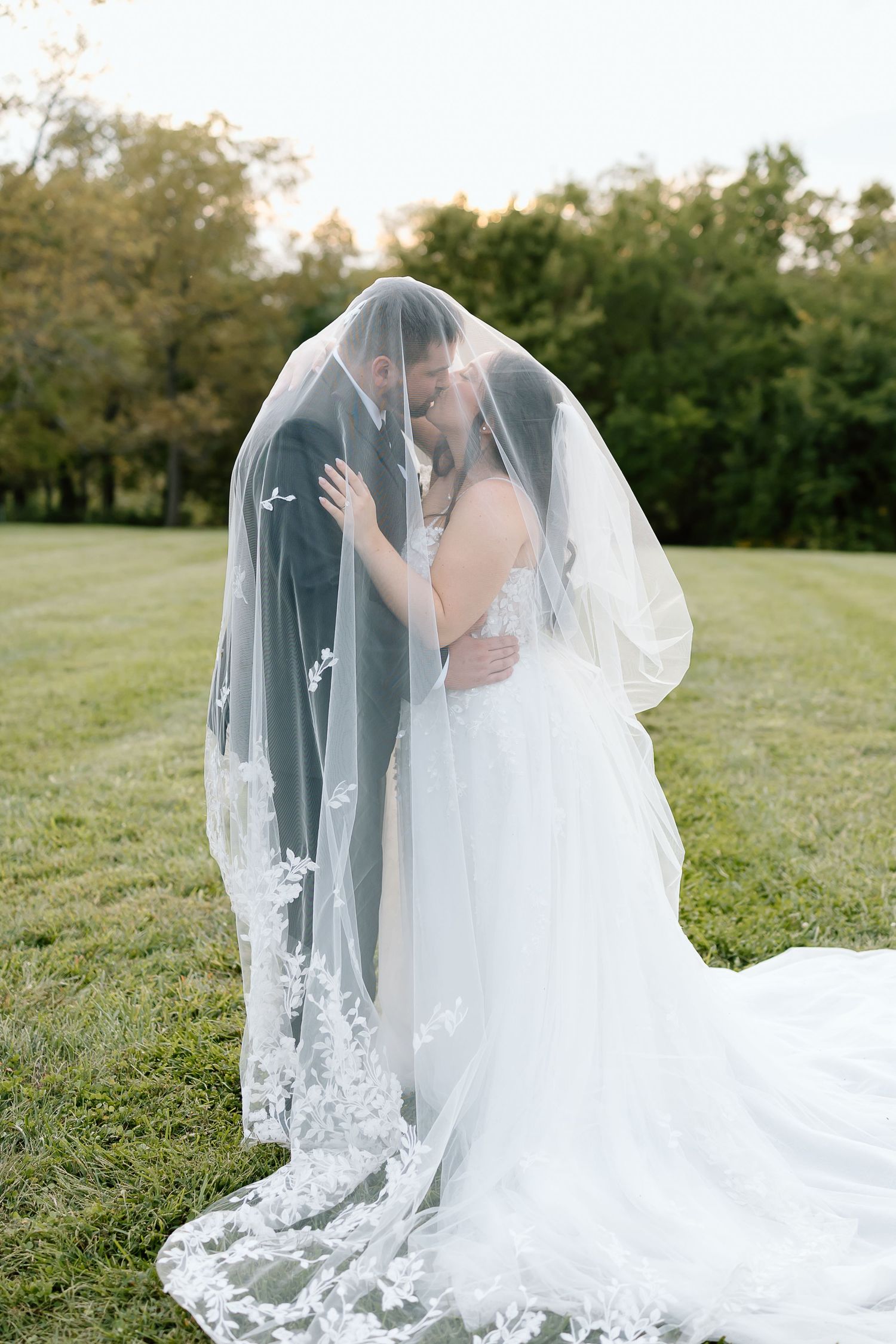 A romantic wedding moment captured outdoors with a flowing lace veil covering the couple beneath trees.