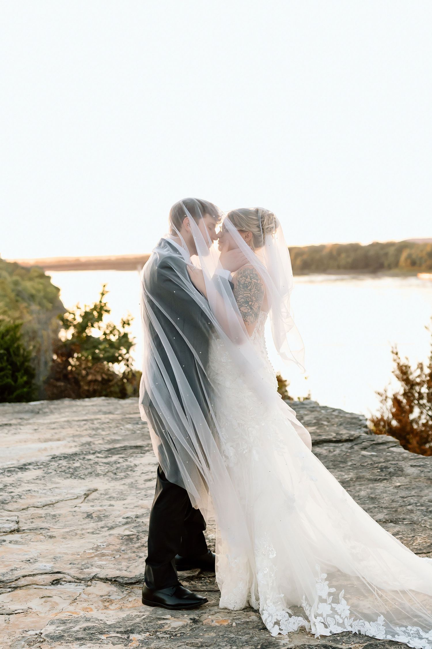 Romantic sunset wedding photos showing a couple embracing on a rocky beach with flowing white veil and gown.