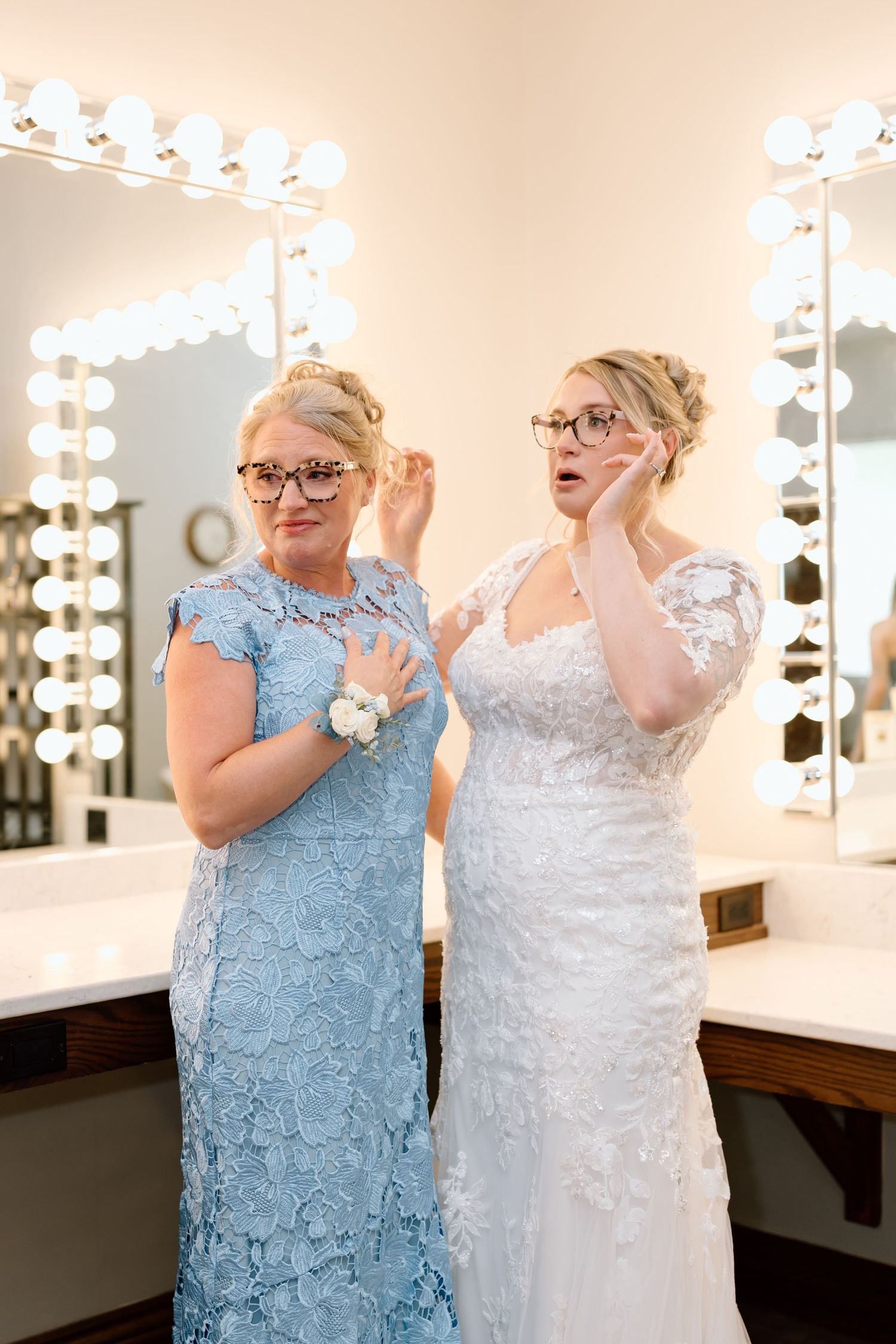 Two women in formal dresses, one in light blue lace and one in white, share a joyful moment in a dressing room.
