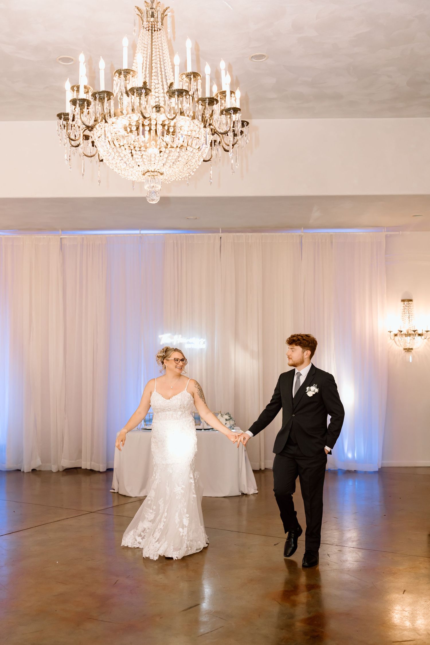 A couple in wedding attire dances across a white ballroom with elegant crystal chandeliers overhead.