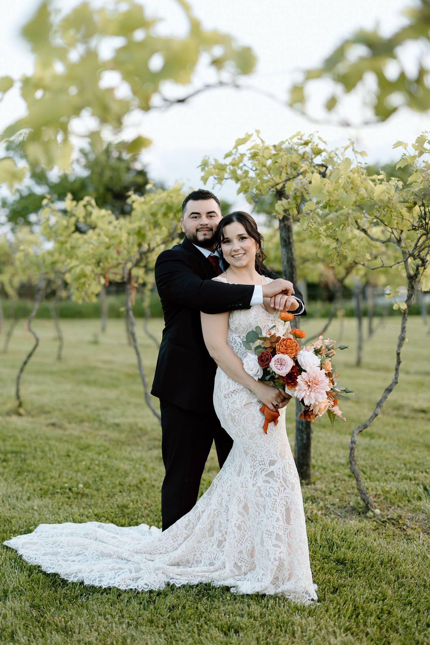 A couple poses playfully in an orchard setting wearing wedding attire with a flowing lace dress and black suit.