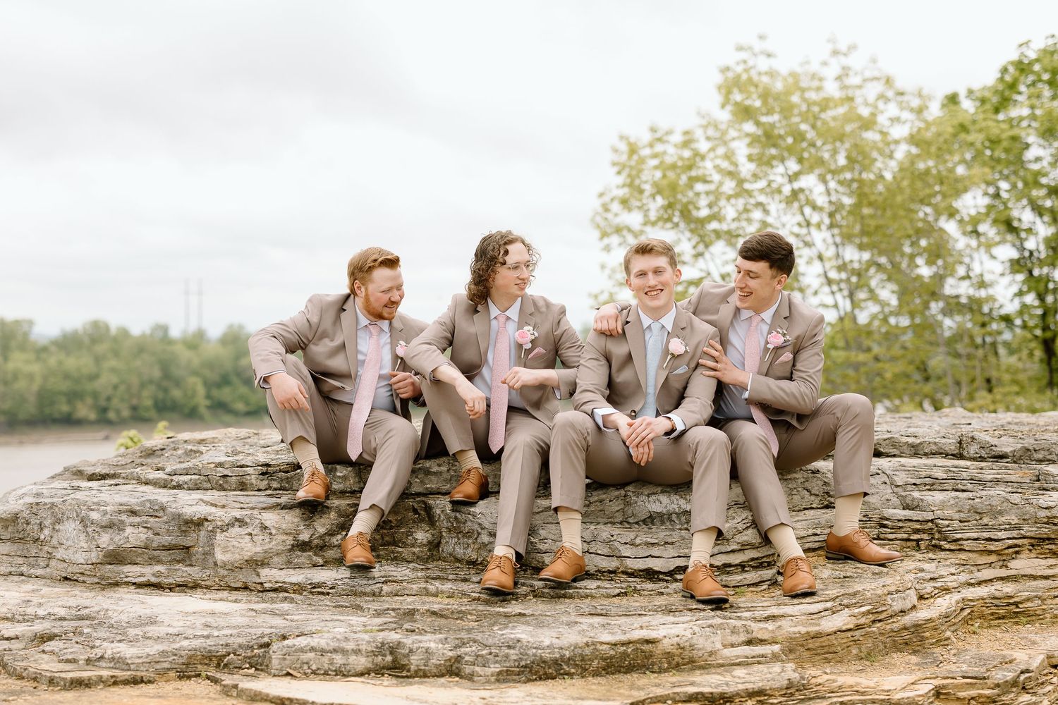 Wedding party in light gray suits relaxes on rustic stone wall during outdoor photo session.