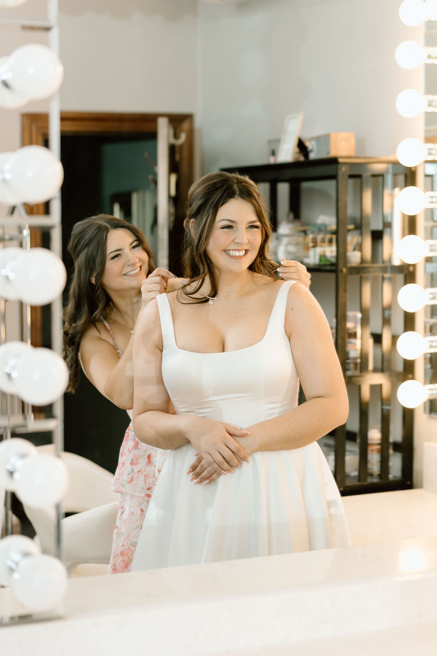 Two women in white outfits smile and embrace while posing in front of a mirror with vanity lights.