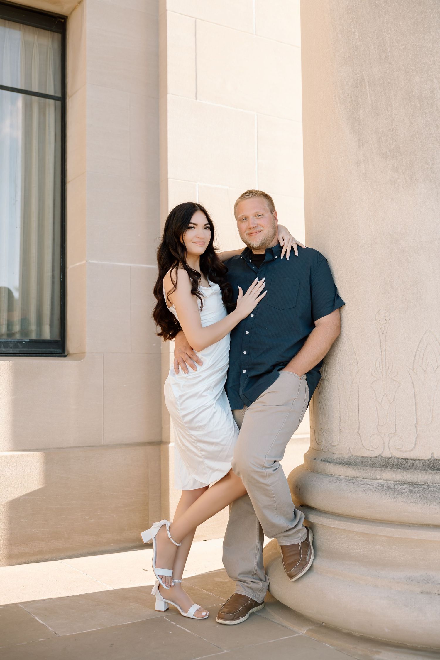 A couple poses playfully against a light colored building wall in a series of romantic engagement photos.
