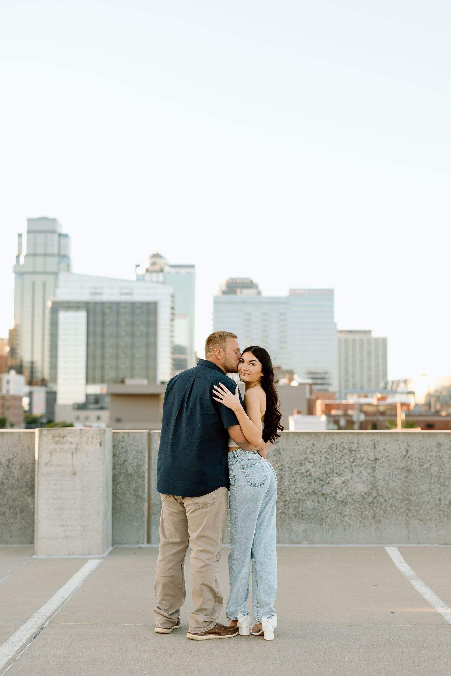 A romantic couple shares tender moments on a rooftop parking deck with a city skyline backdrop.