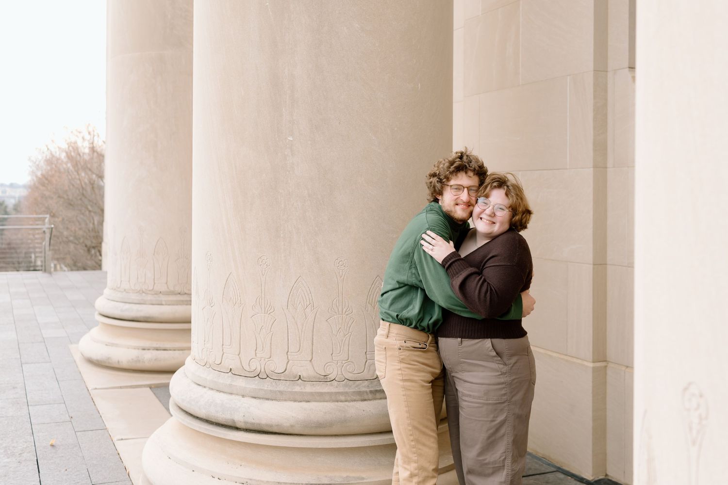 A couple embraces and shares a romantic moment between elegant white stone columns in a classical architectural setting.