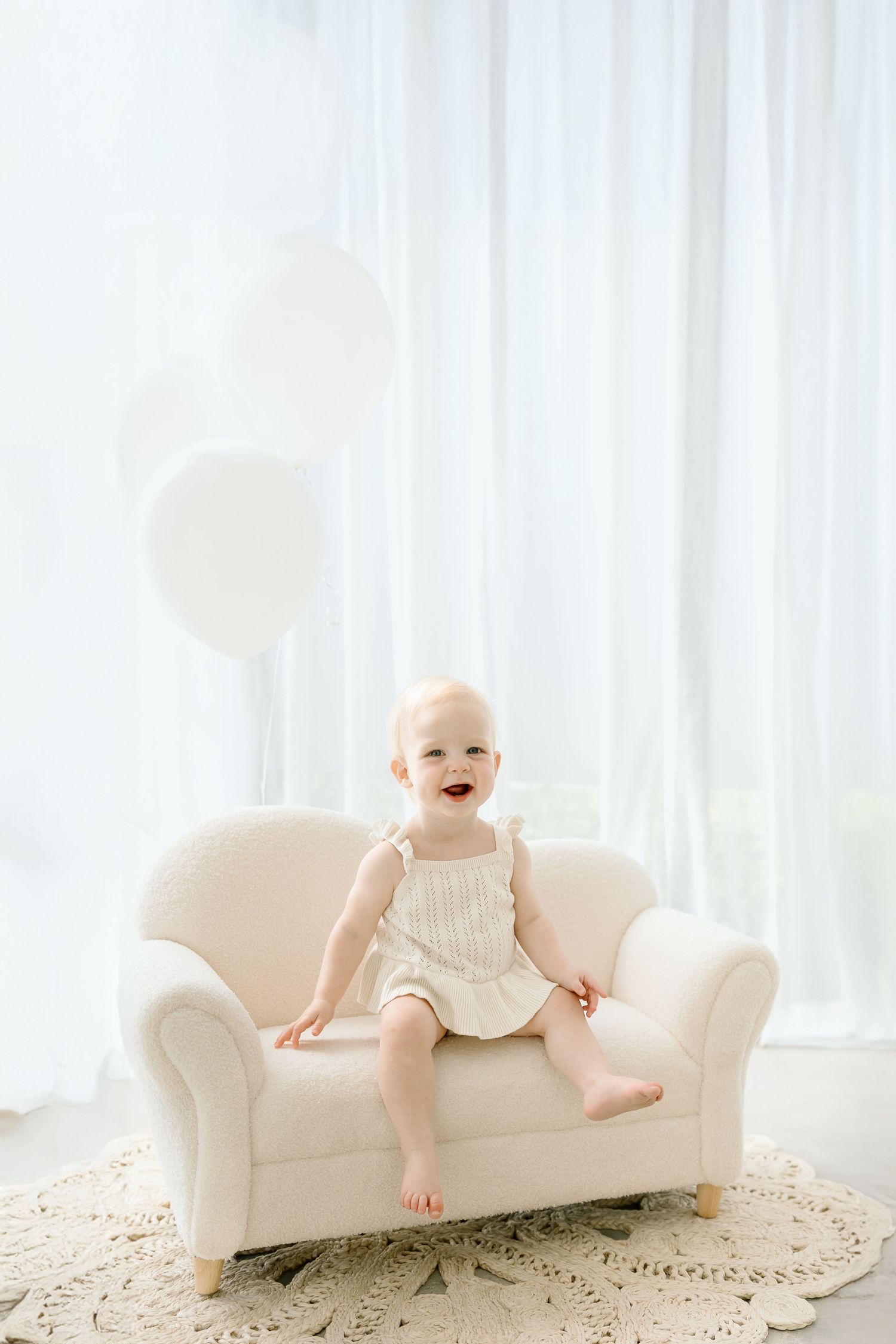 A happy baby in white romper sits and plays on a white couch in a bright, airy room.