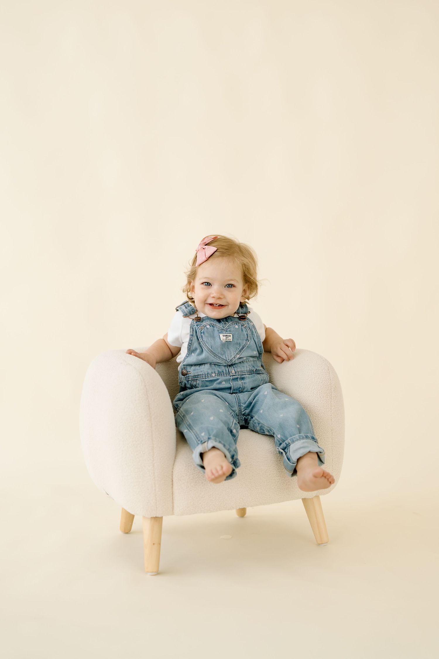 A happy baby in denim overalls sits and smiles while lounging in a modern white chair against a neutral background.