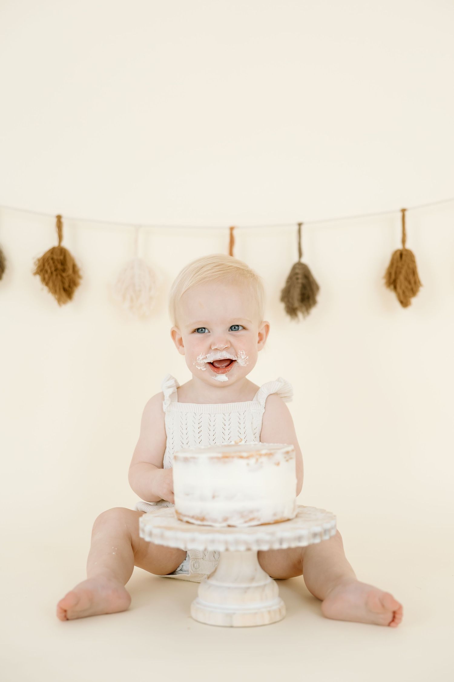 A baby in white sits with a white birthday cake against a light neutral background.