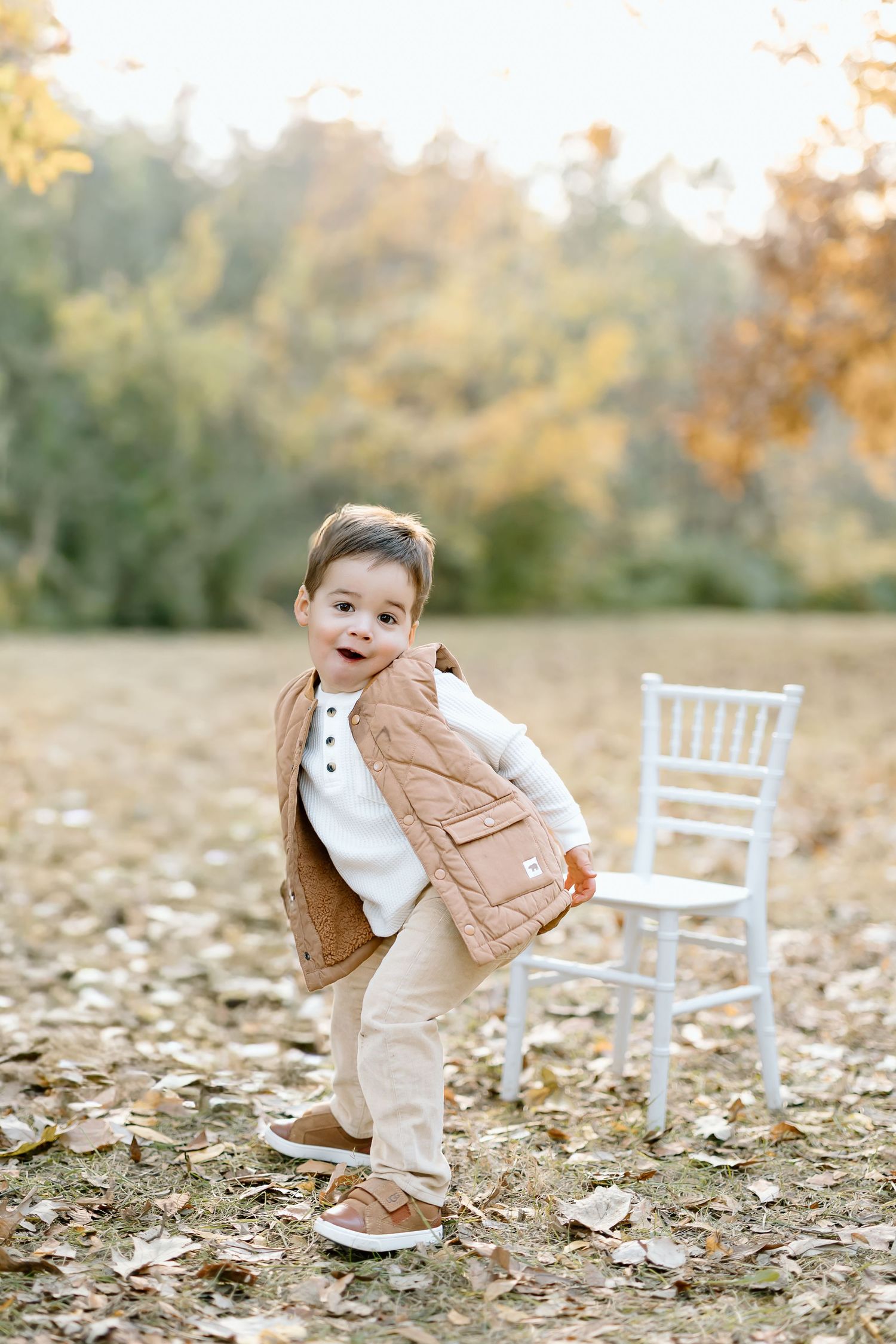 A young child in a tan vest and white shirt plays in autumn leaves near white chairs in an outdoor setting.