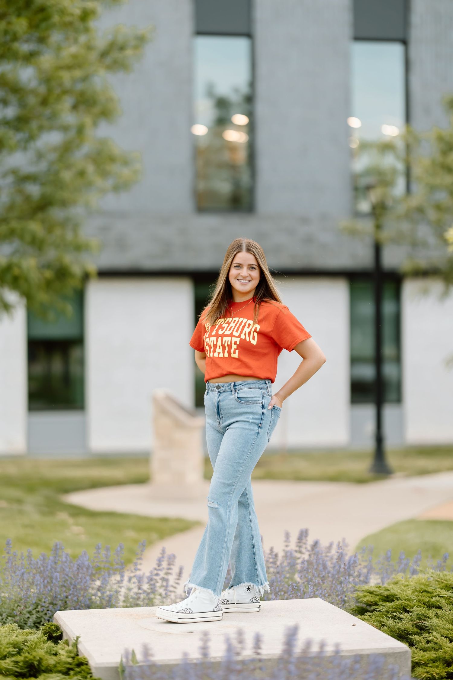 A person in an orange t-shirt and light blue jeans poses outdoors near purple flowers and modern architecture.