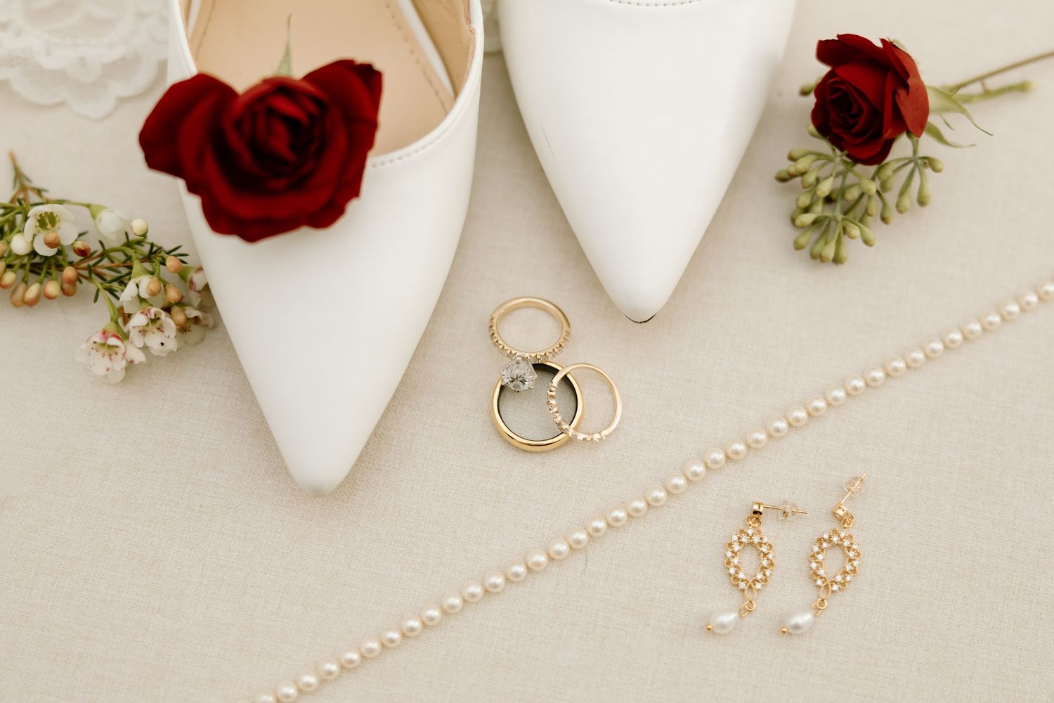 Wedding accessories including white heels, red roses, pearl necklace, gold earrings and engagement ring on beige background.