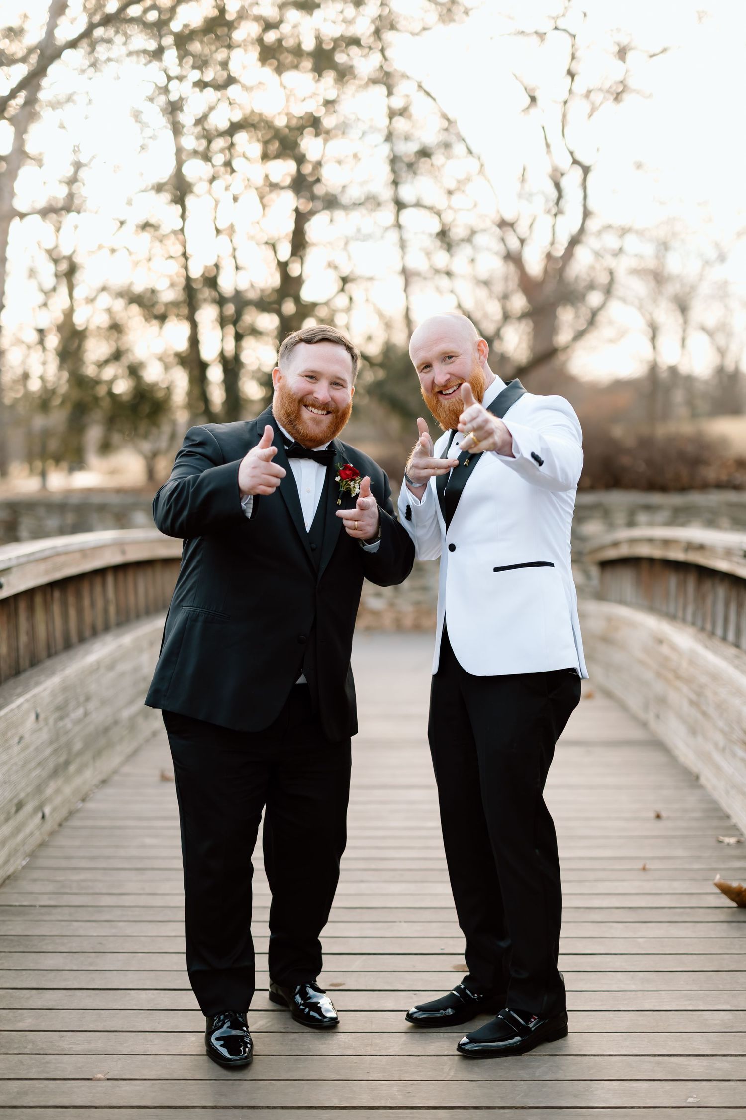Two people in formal black and white attire pose playfully on a wooden bridge during a wedding photoshoot.