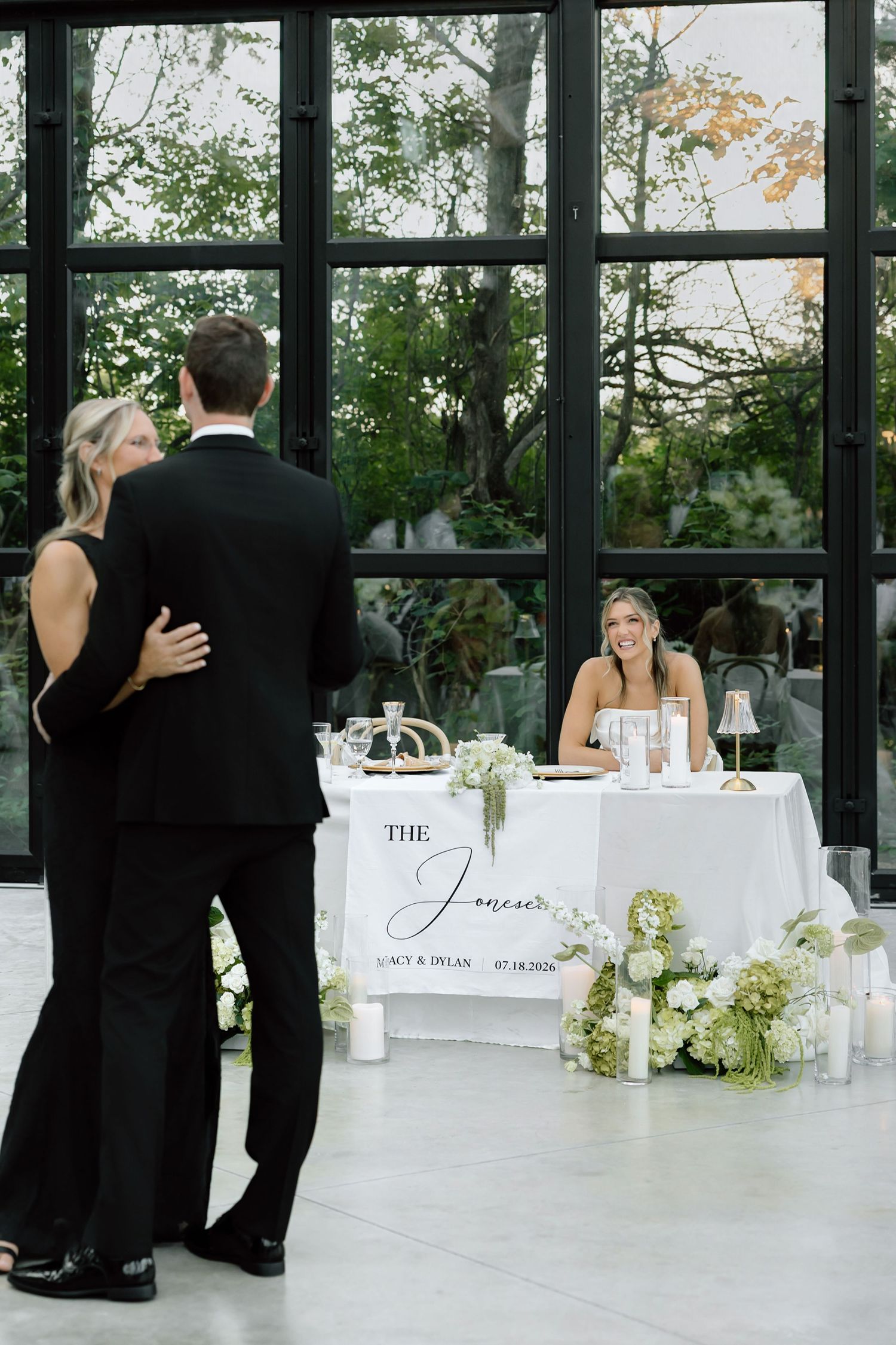 Couple dancing at wedding reception near sweetheart table with floral arrangements and large black-framed windows.