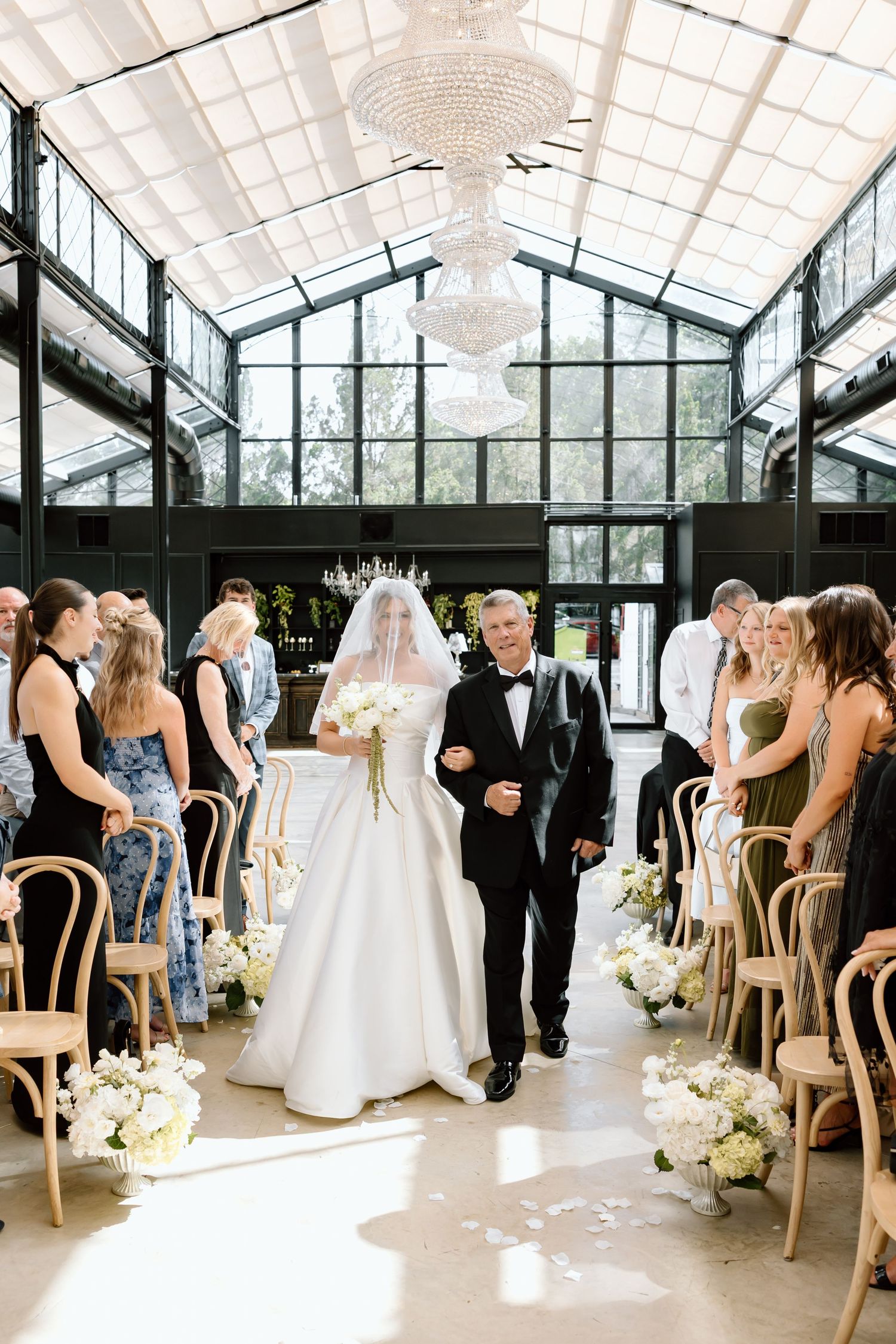 Bride walks down the aisle in a greenhouse wedding venue with chandelier, white florals, and bentwood chairs.