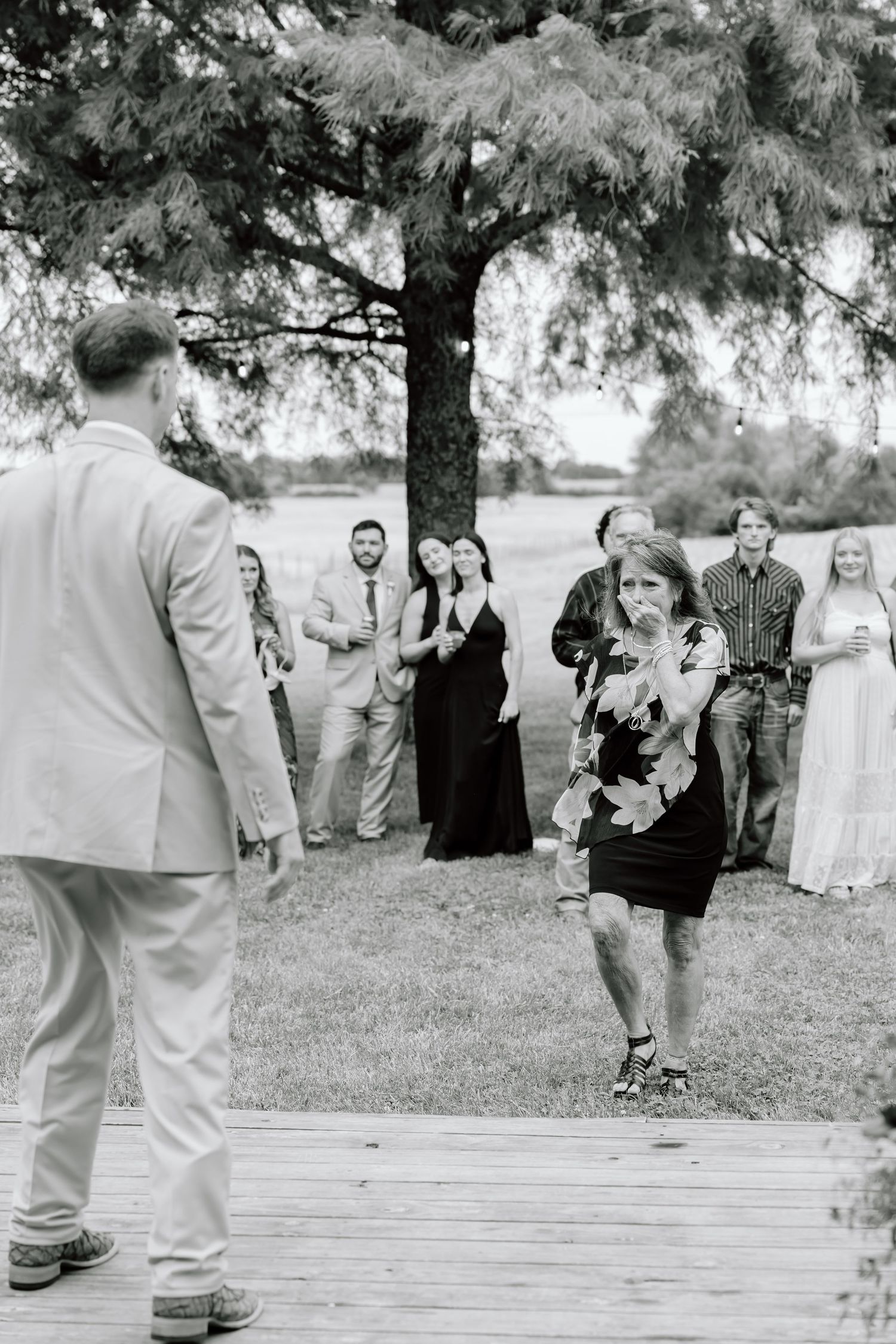 Black and white wedding photo of guests watching a woman walk toward a groom on an outdoor wooden deck.