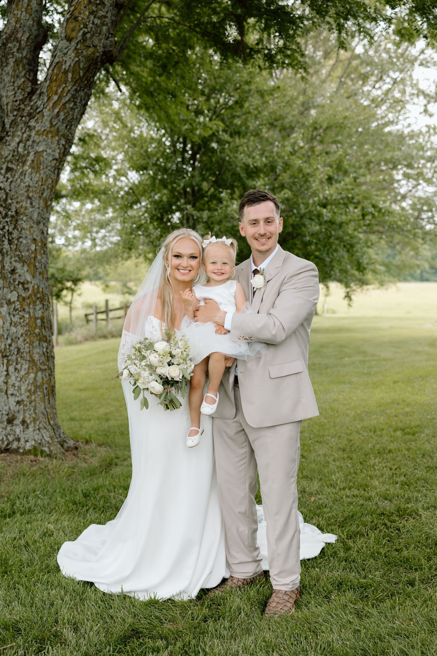 Bride in white gown and groom in tan suit hold toddler girl outdoors under large tree on lush green lawn.