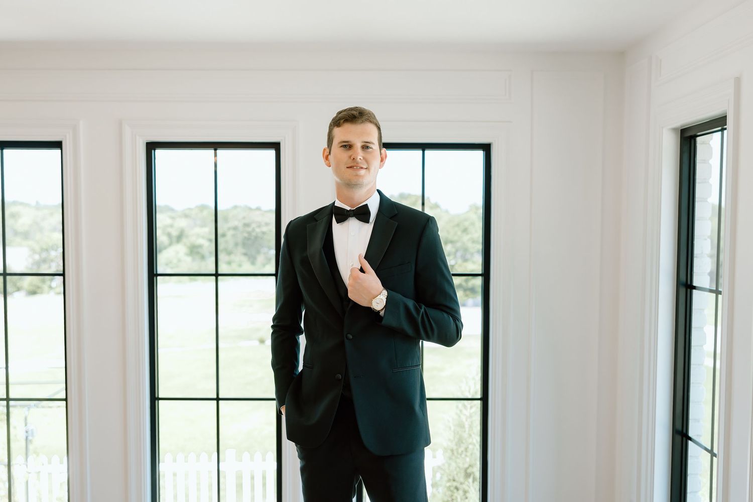 A dapper groom in a black tuxedo with bow tie poses confidently in a bright white room with large windows.
