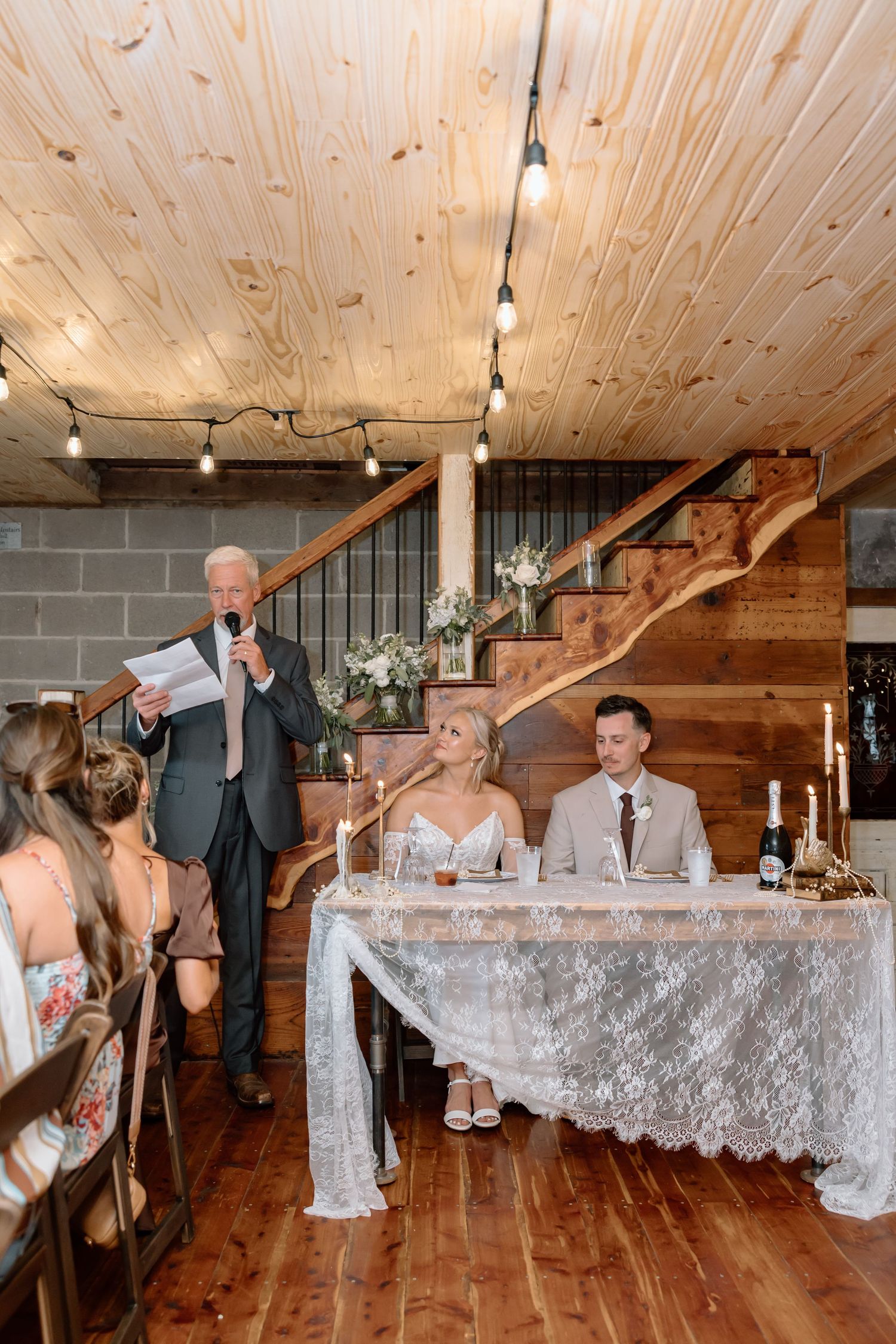 Elderly man giving a wedding toast at a rustic barn reception while bride and groom sit at a lace-covered sweetheart table.