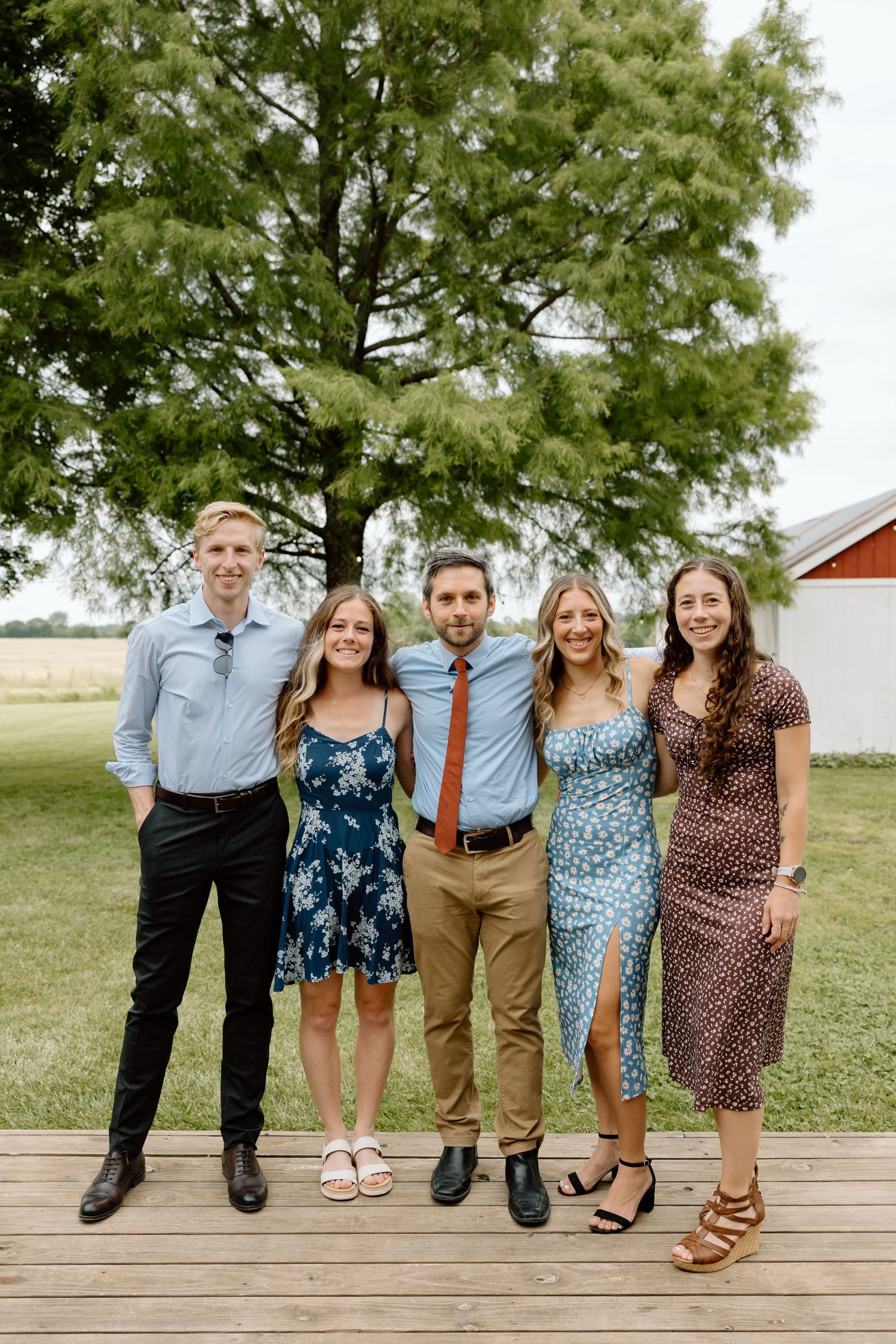 Five young adults smiling together on a wooden deck outdoors near a large tree and red barn.