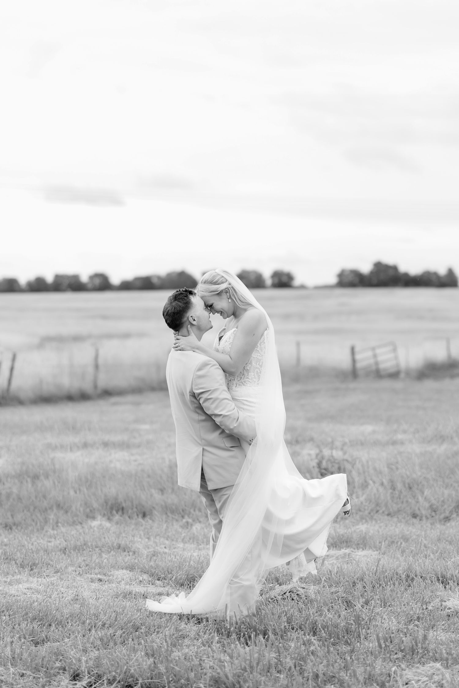 Black and white photo of a groom lifting his bride in a field, sharing a romantic kiss on their wedding day.