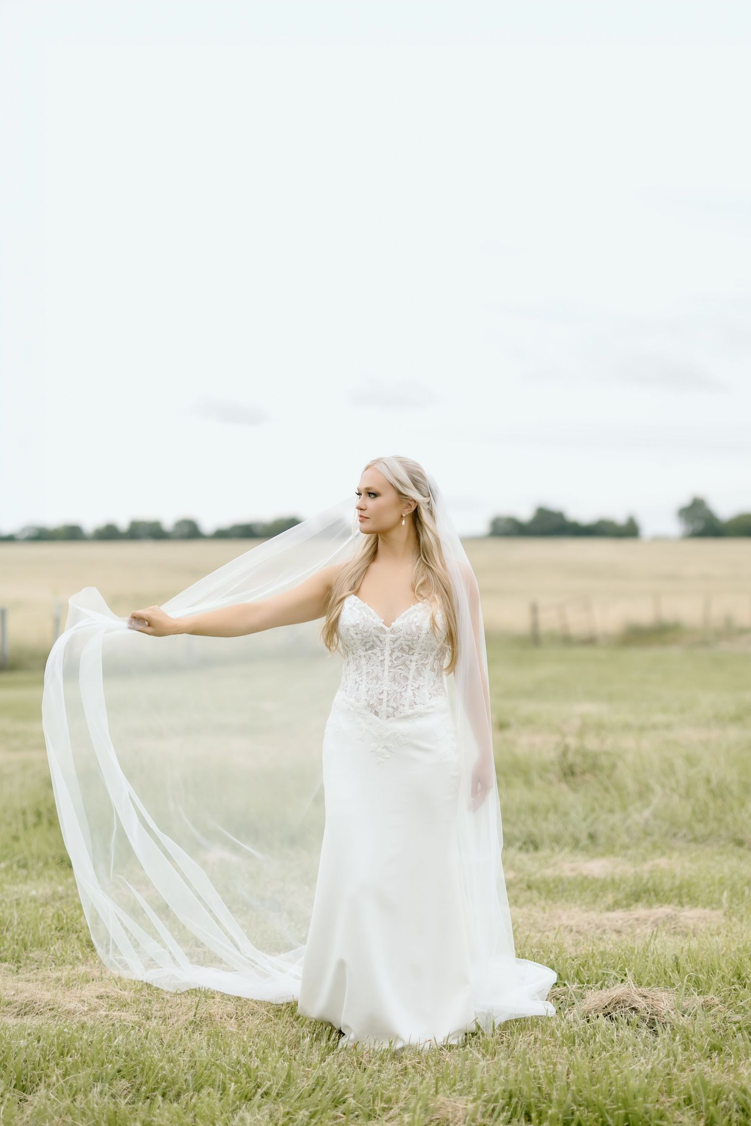 Bride in white lace corset gown holding flowing veil in green field on overcast day.