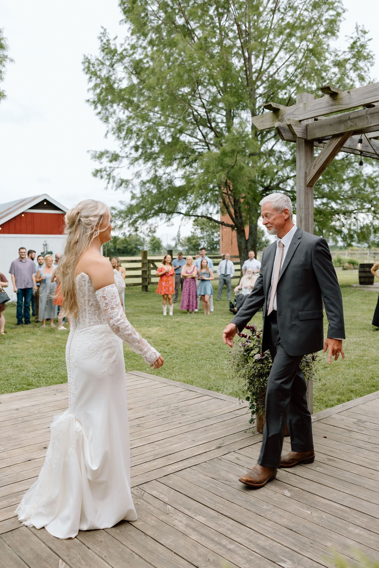 Bride in lace wedding gown walks with older man in gray suit on wooden deck at outdoor farm wedding ceremony.