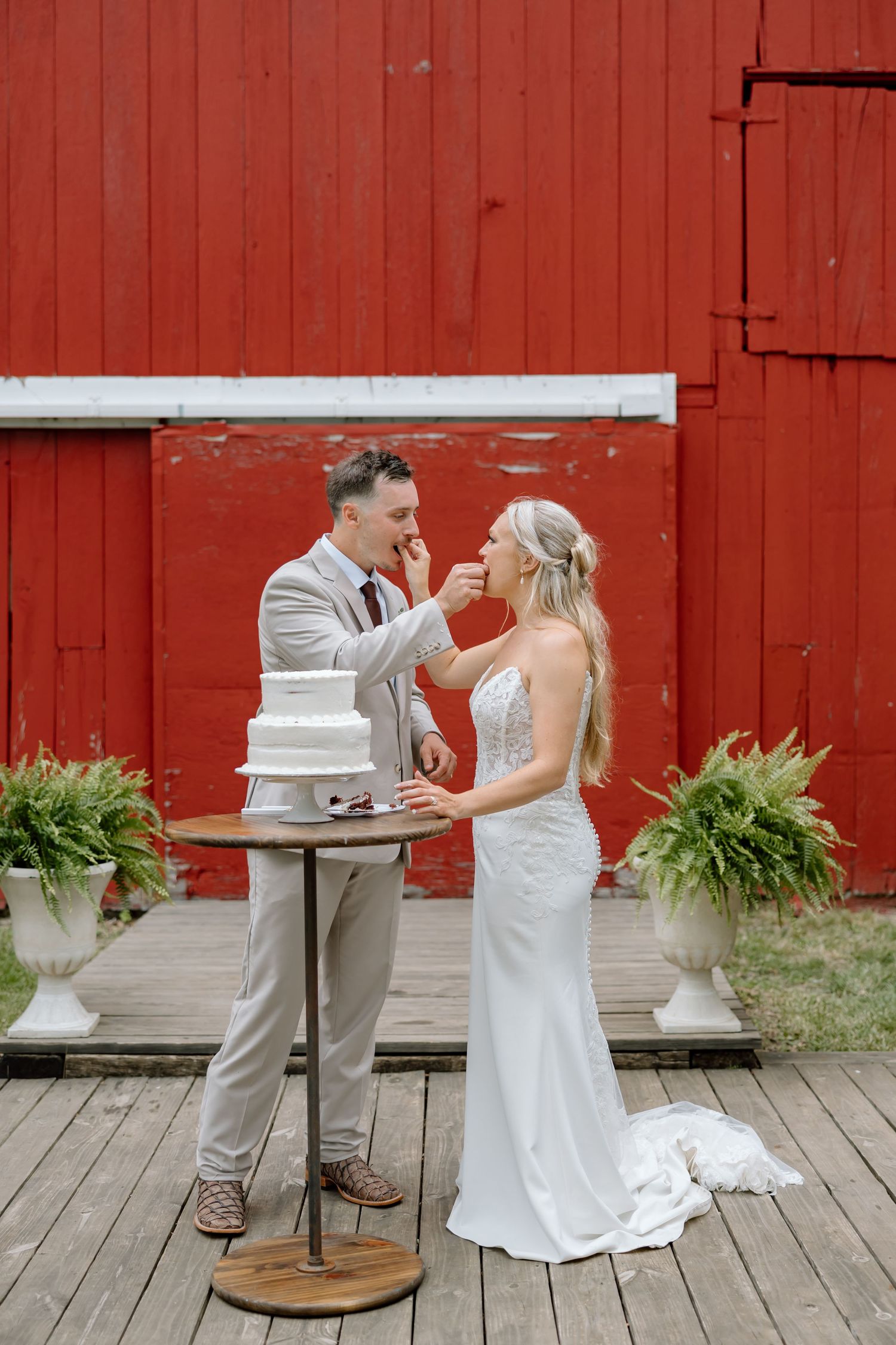 Bride and groom feeding each other wedding cake on a rustic wooden deck in front of a red barn.