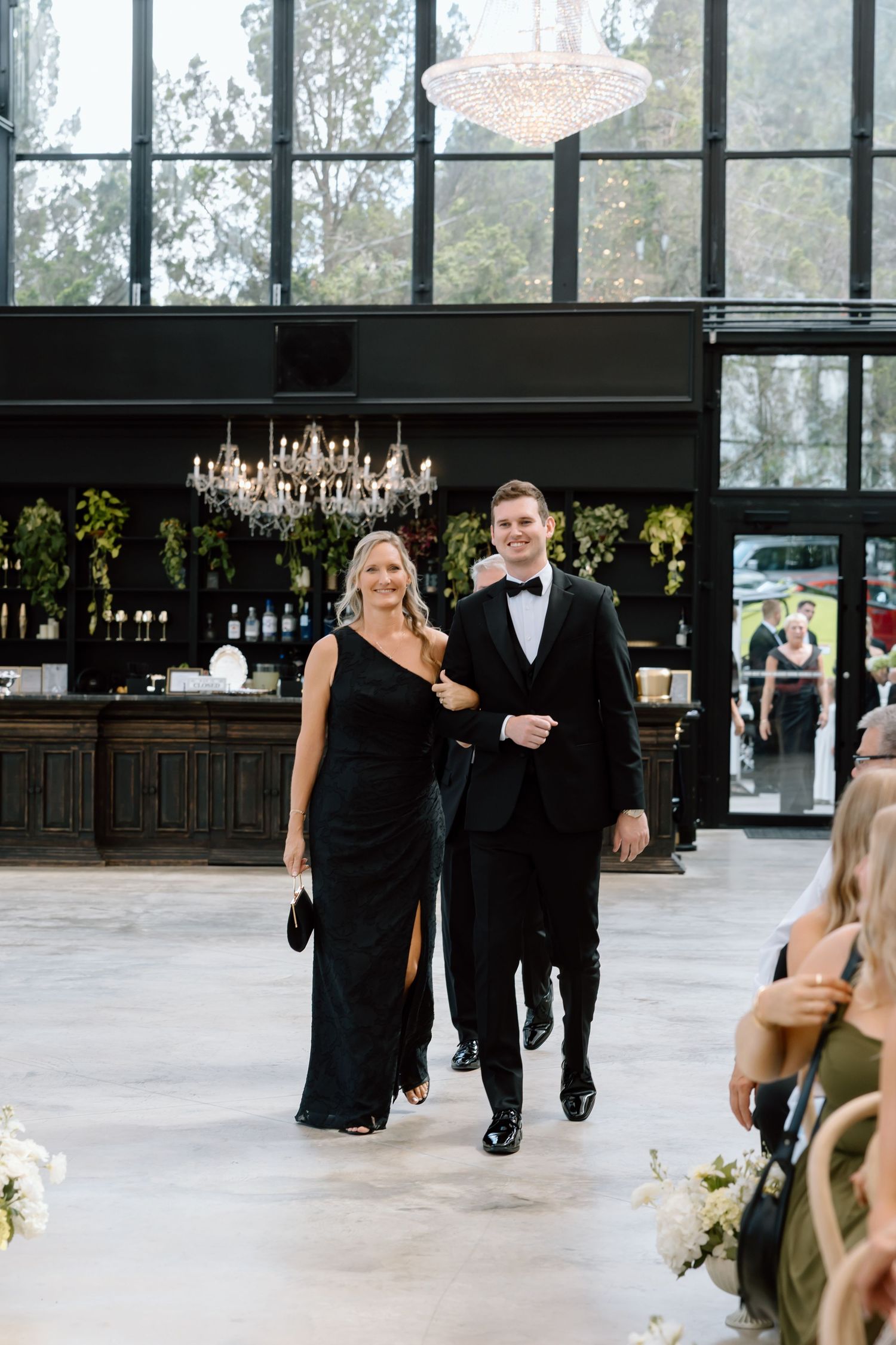 A woman in a black gown and a man in a tuxedo walk arm-in-arm through an elegant venue with chandeliers.