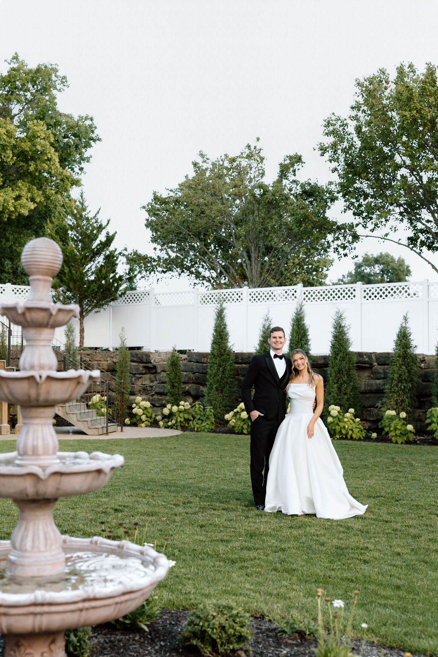 Bride and groom pose elegantly in a garden with a stone fountain, lush greenery, and white lattice fence backdrop.