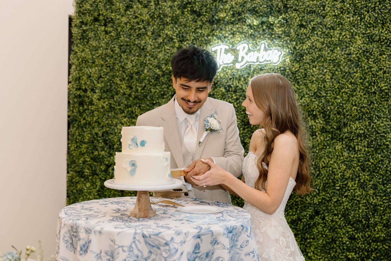 Bride and groom cutting white wedding cake at reception with green hedge wall backdrop.