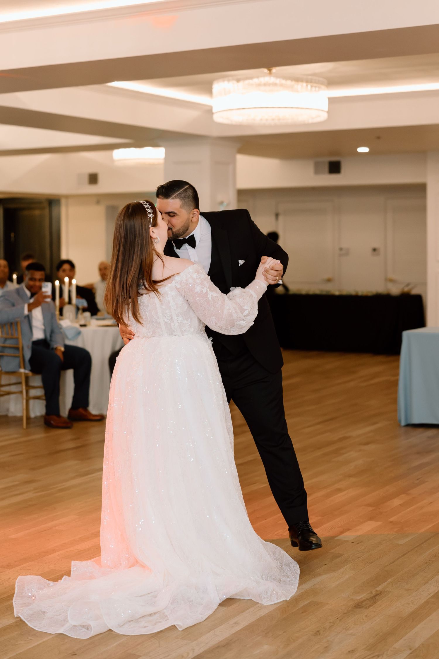 Bride and groom share their first dance in a elegant ballroom, groom in black tuxedo kissing bride in lace gown.