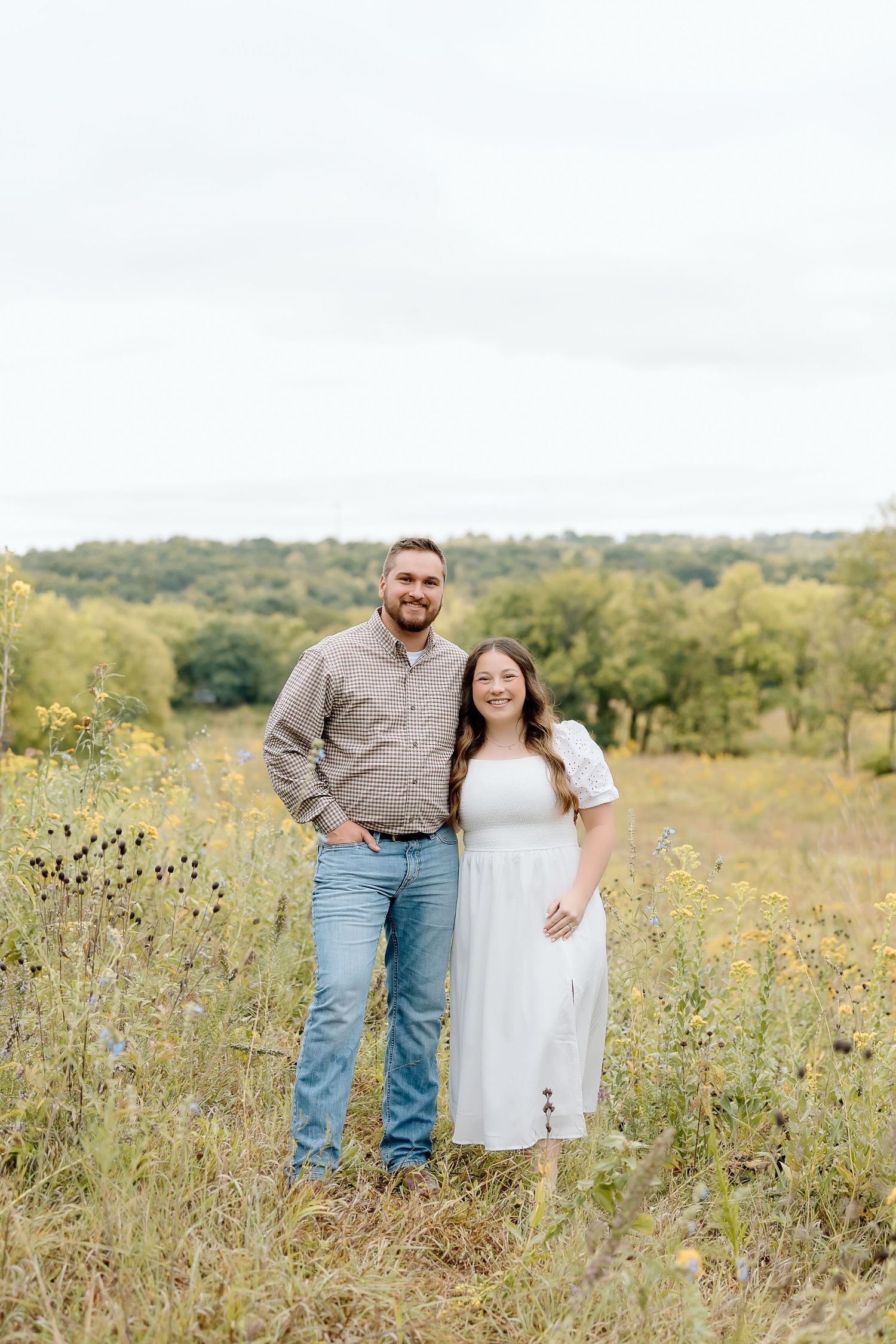 Happy couple poses in a wildflower meadow with trees and hills in the background.